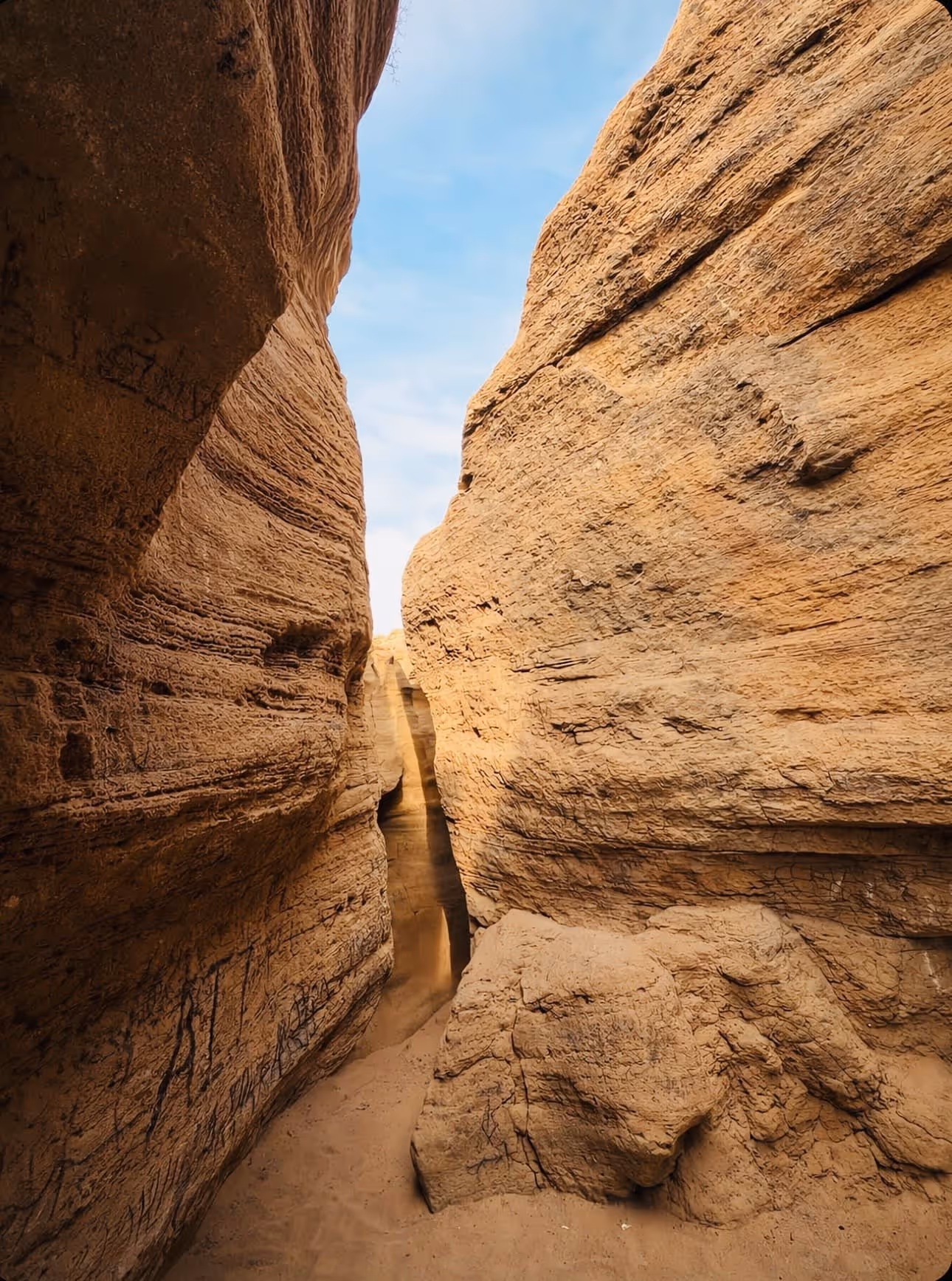 Narrow sandy path between tall textured sandstone canyon walls under a blue sky.