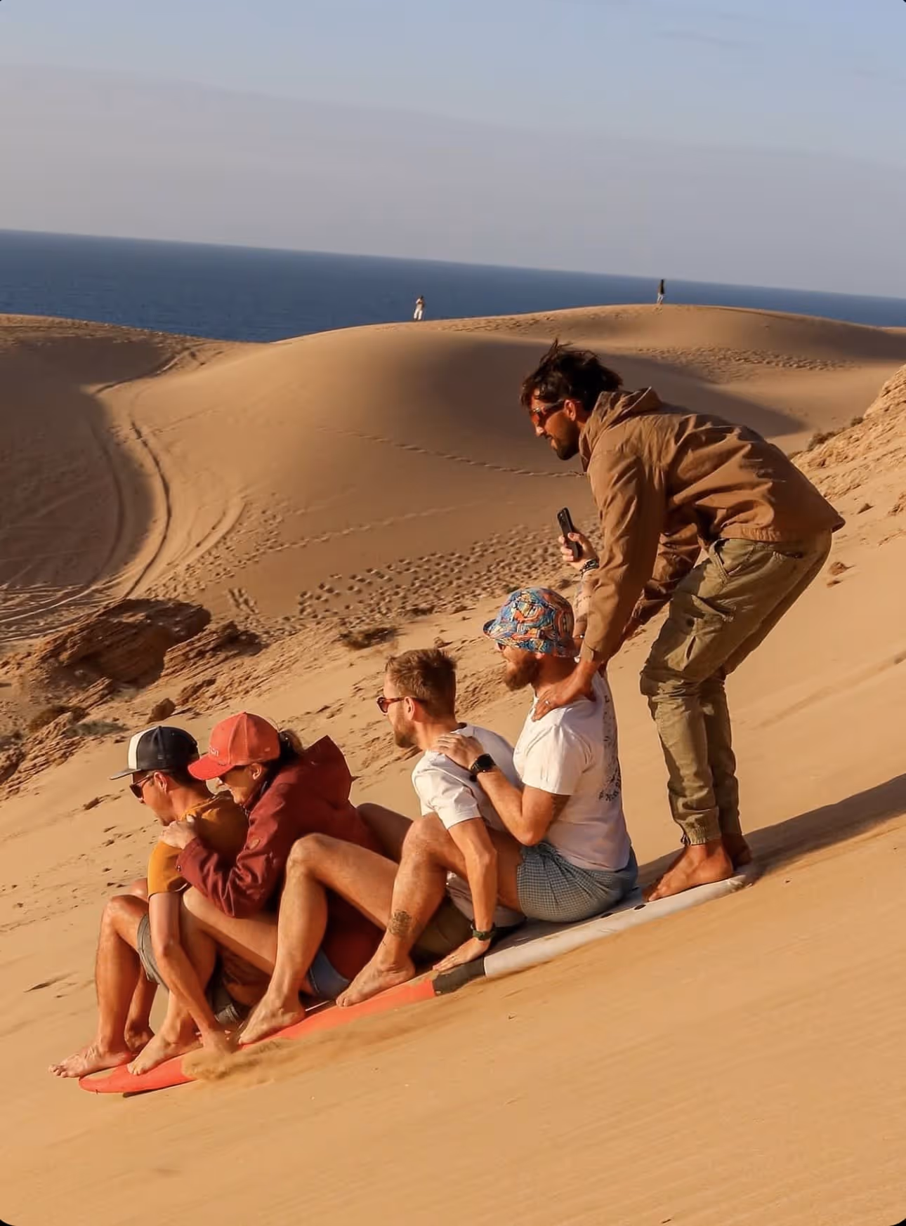 Five people sandboarding together on a desert dune near the ocean with two standing in the background.
