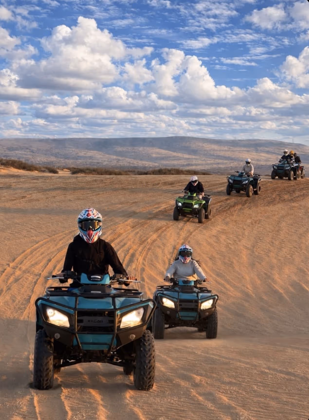 Seven people riding all-terrain vehicles on sandy terrain under a partly cloudy sky.
