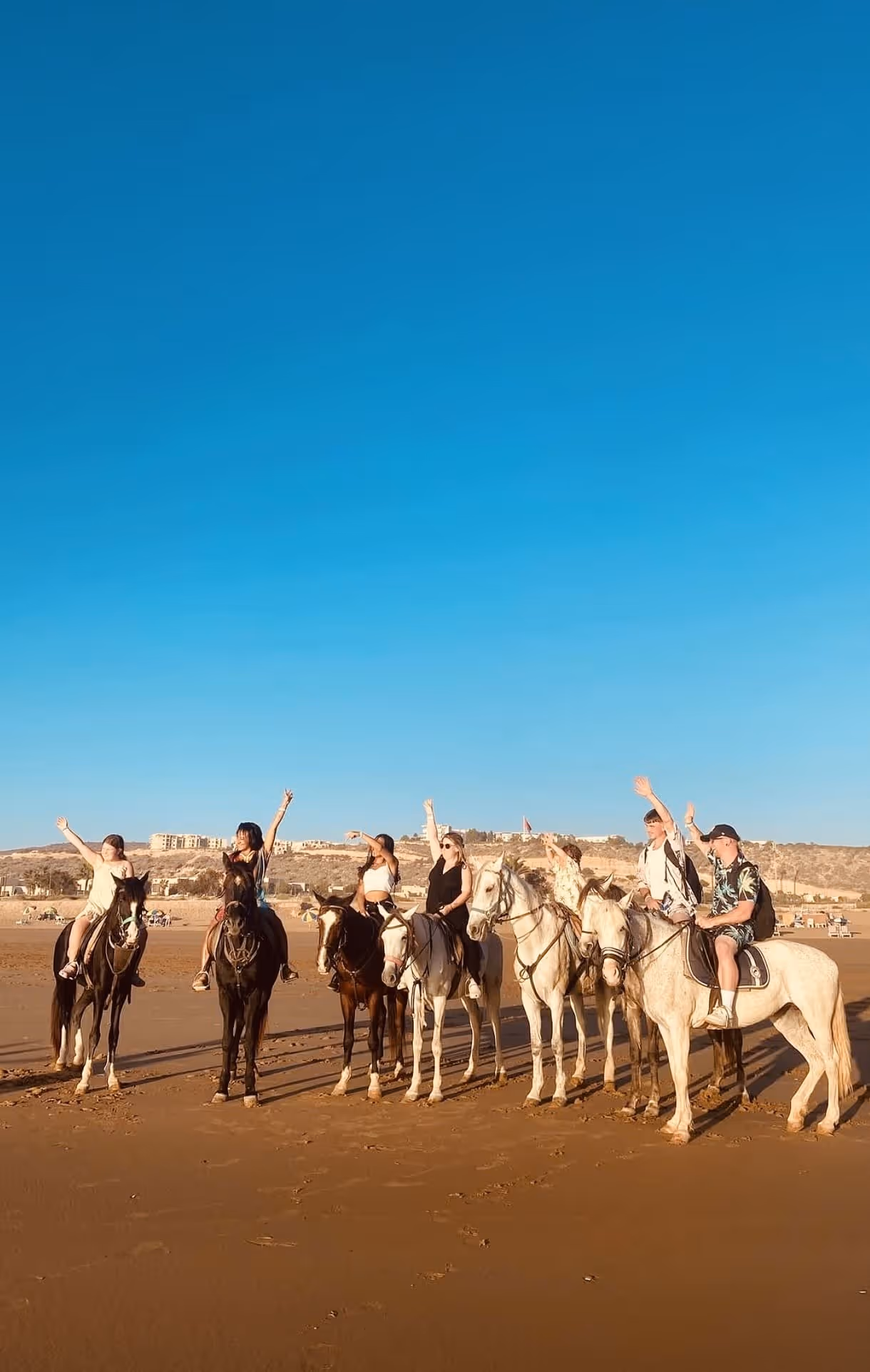 Group of seven people riding horses on a beach under a clear blue sky, waving their hands.