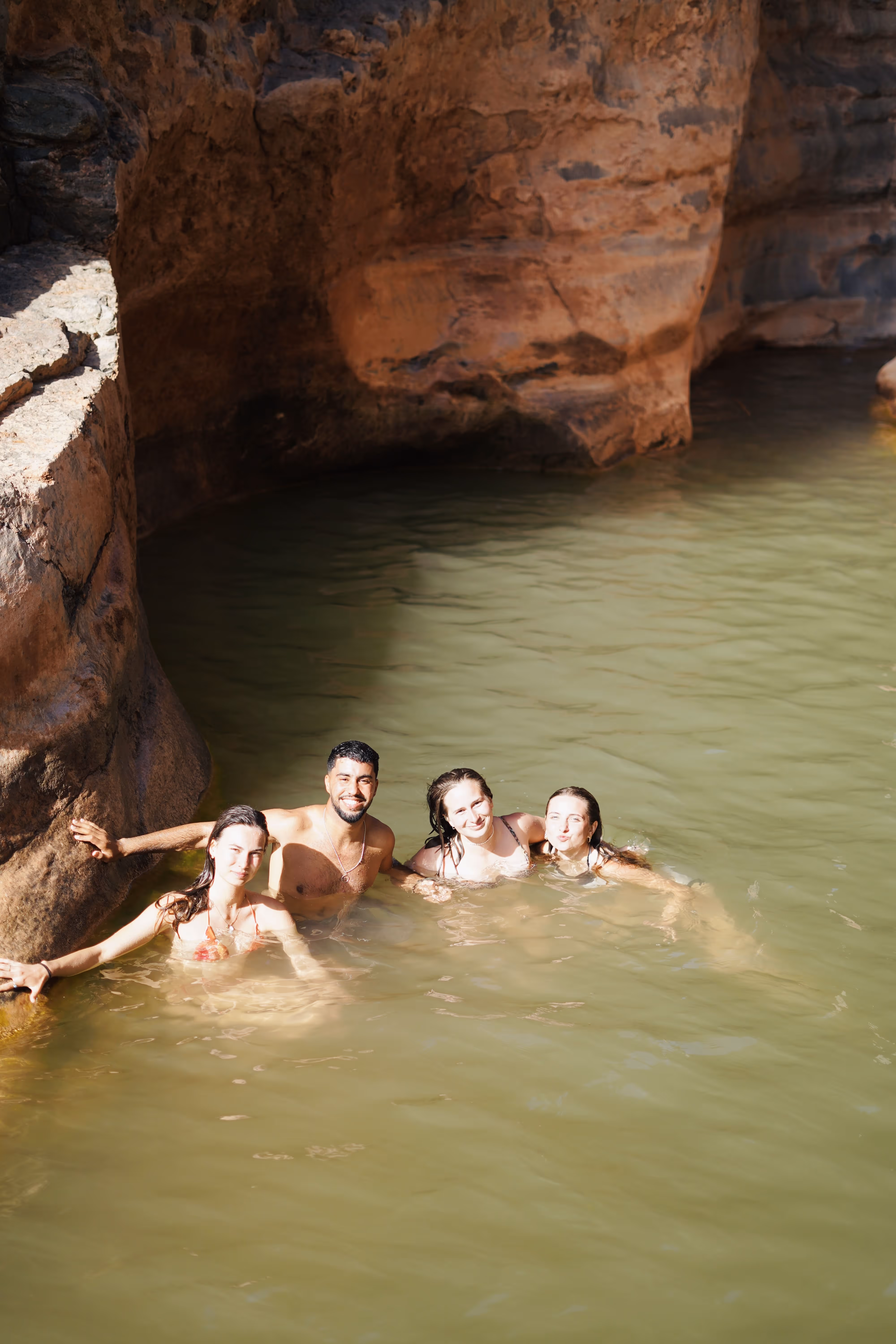 Four young people swimming and smiling in a natural rock pool with greenish water.