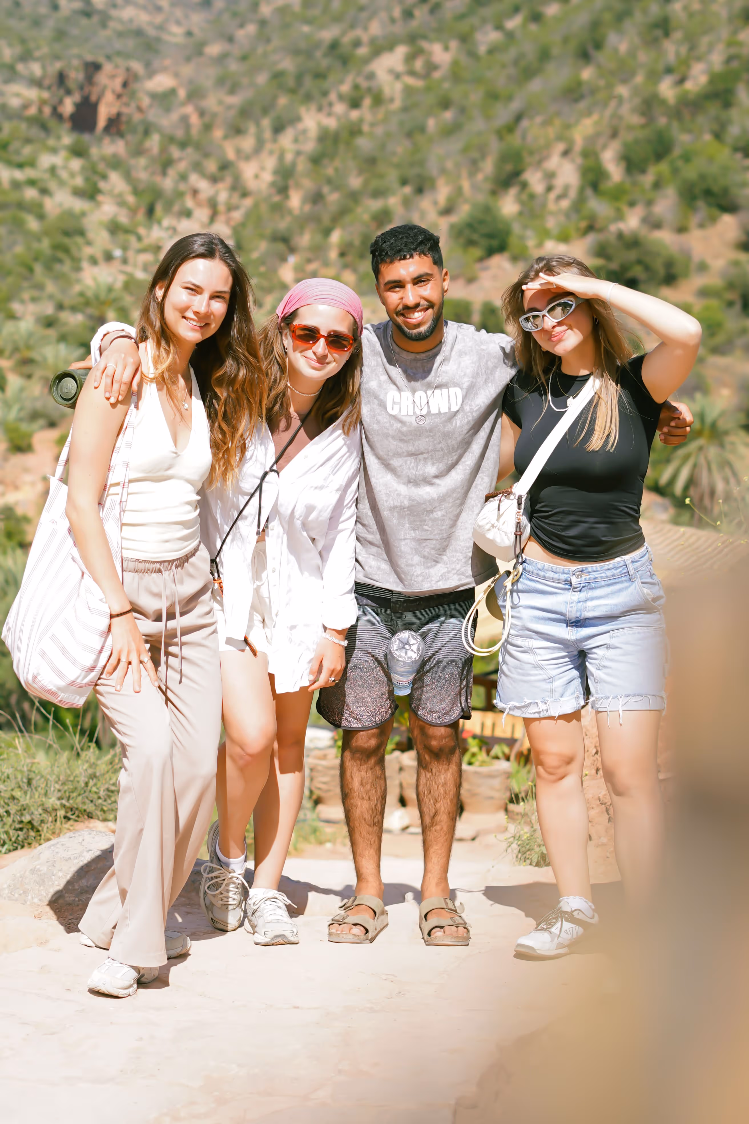 Four friends standing outdoors with arms around each other, smiling, mountains in background.