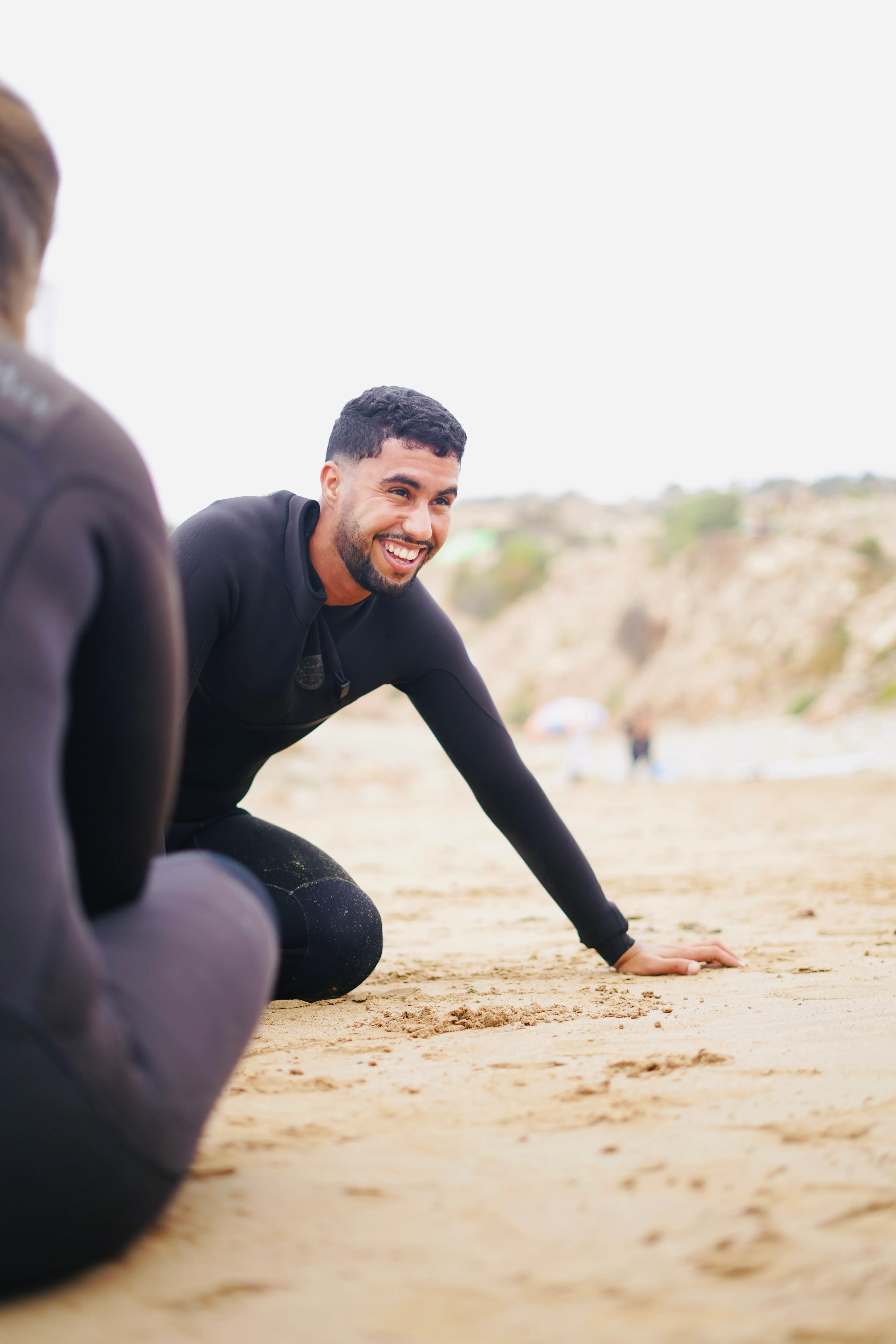 Smiling man in a wetsuit kneeling on sandy beach near another person also in a wetsuit.