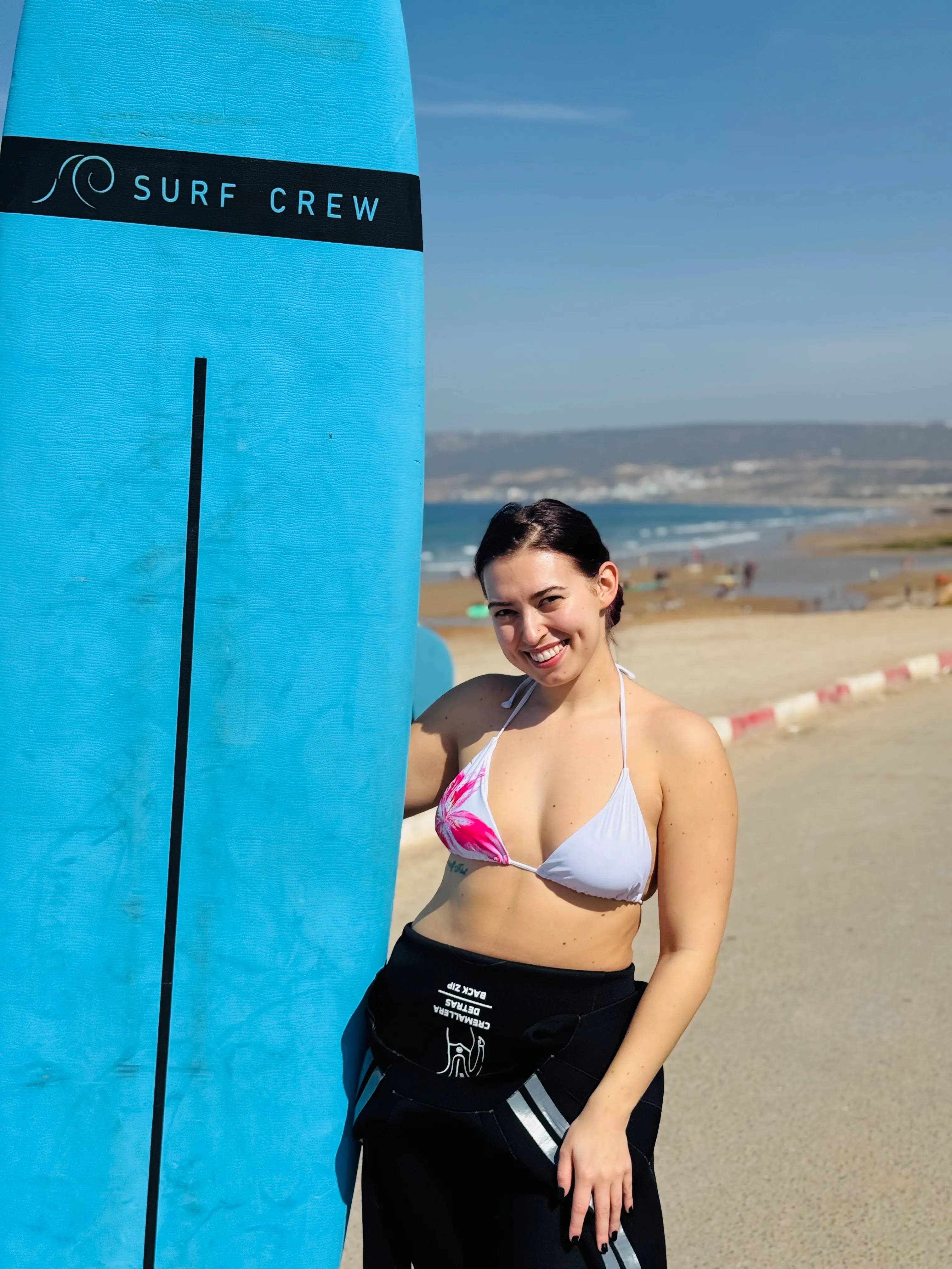 Smiling woman in bikini top and wetsuit holding blue surfboard marked 'Surf Crew' on sunny beach.