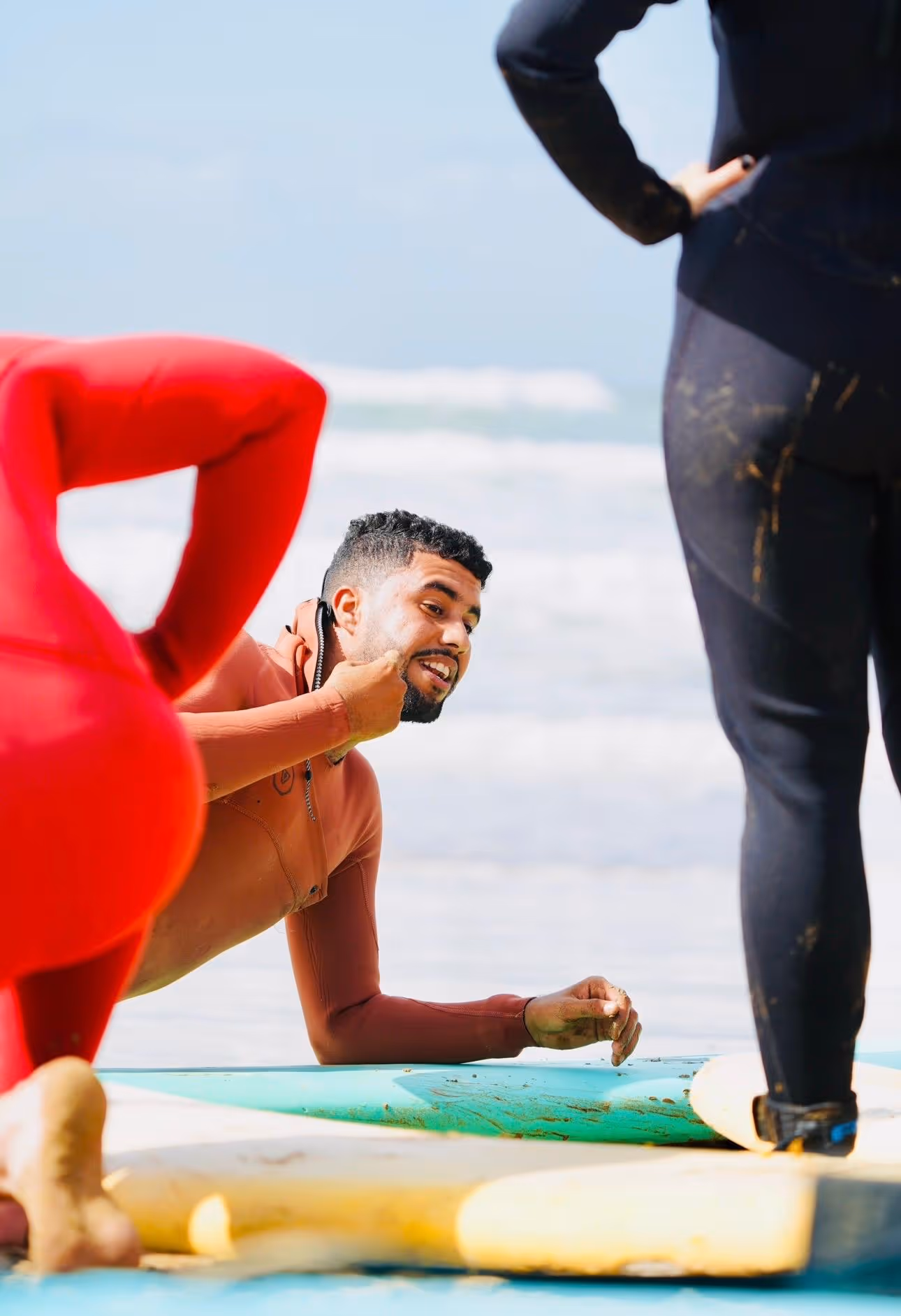Man in wetsuit demonstrating a surfing technique on the beach with two standing people in wetsuits.
