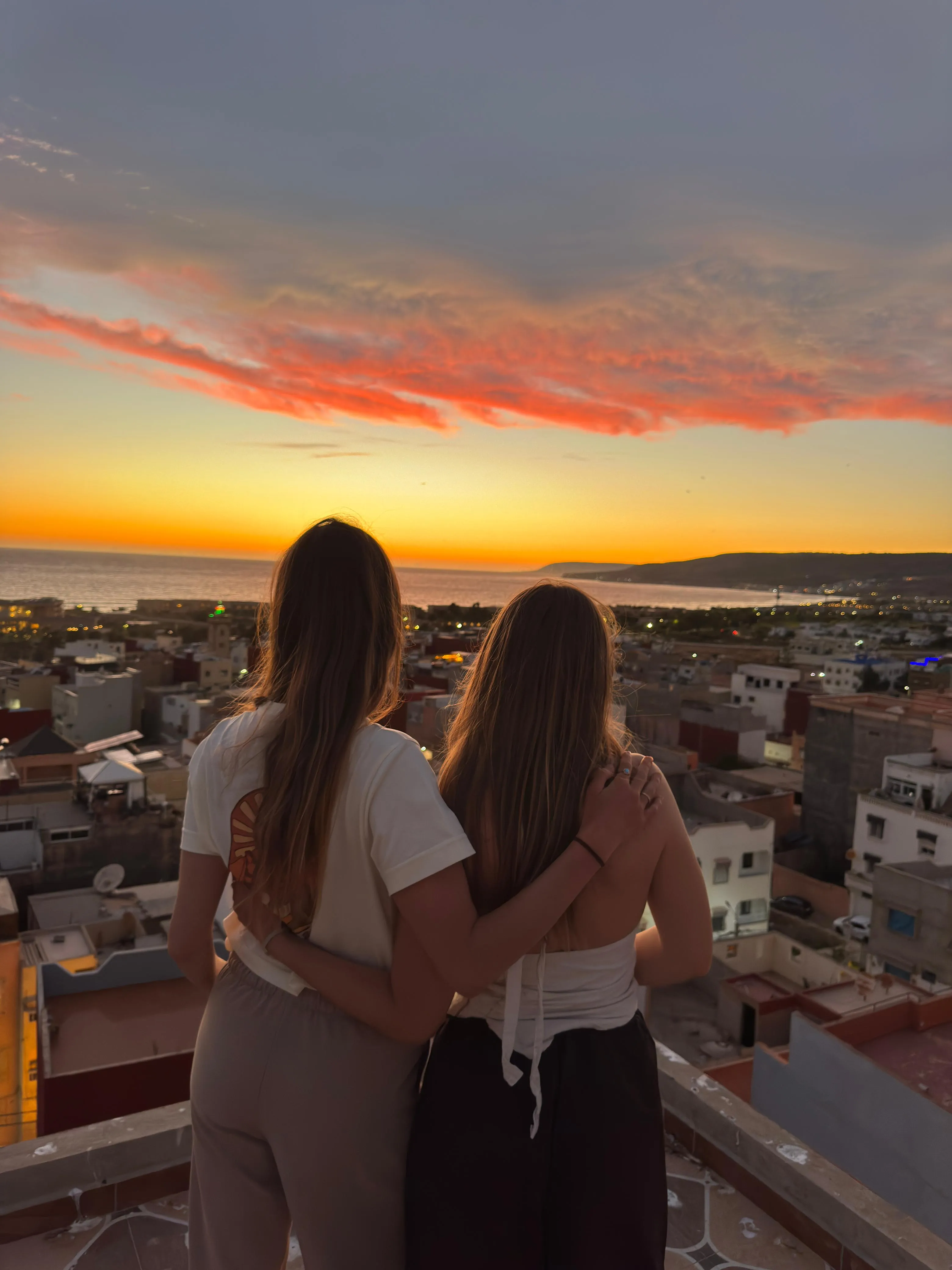 Two women hugging on rooftop overlooking city and sea at sunset with orange and pink clouds.