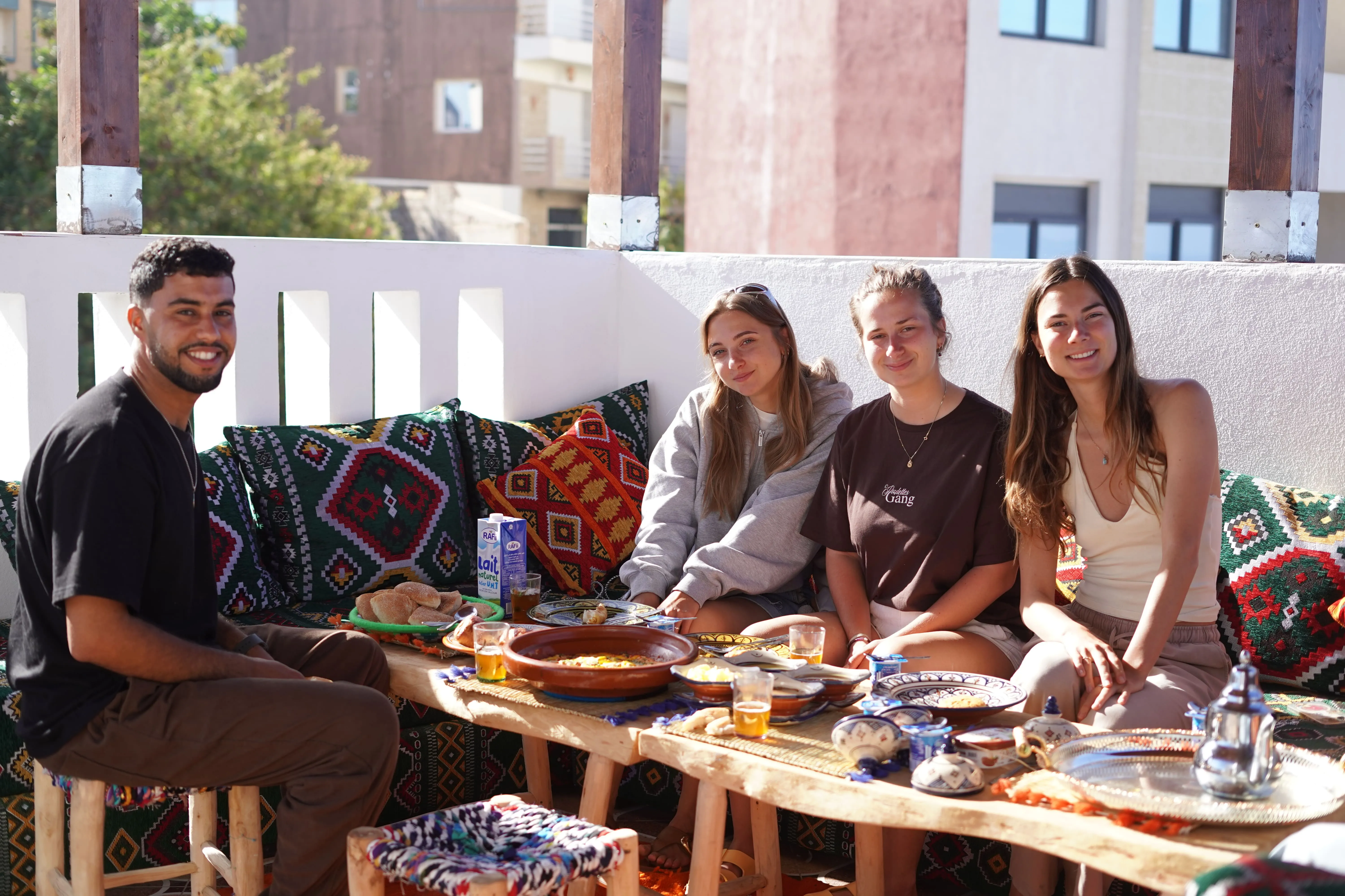 Group of four people sitting on patterned cushions around table with traditional dishes and tea outdoors.