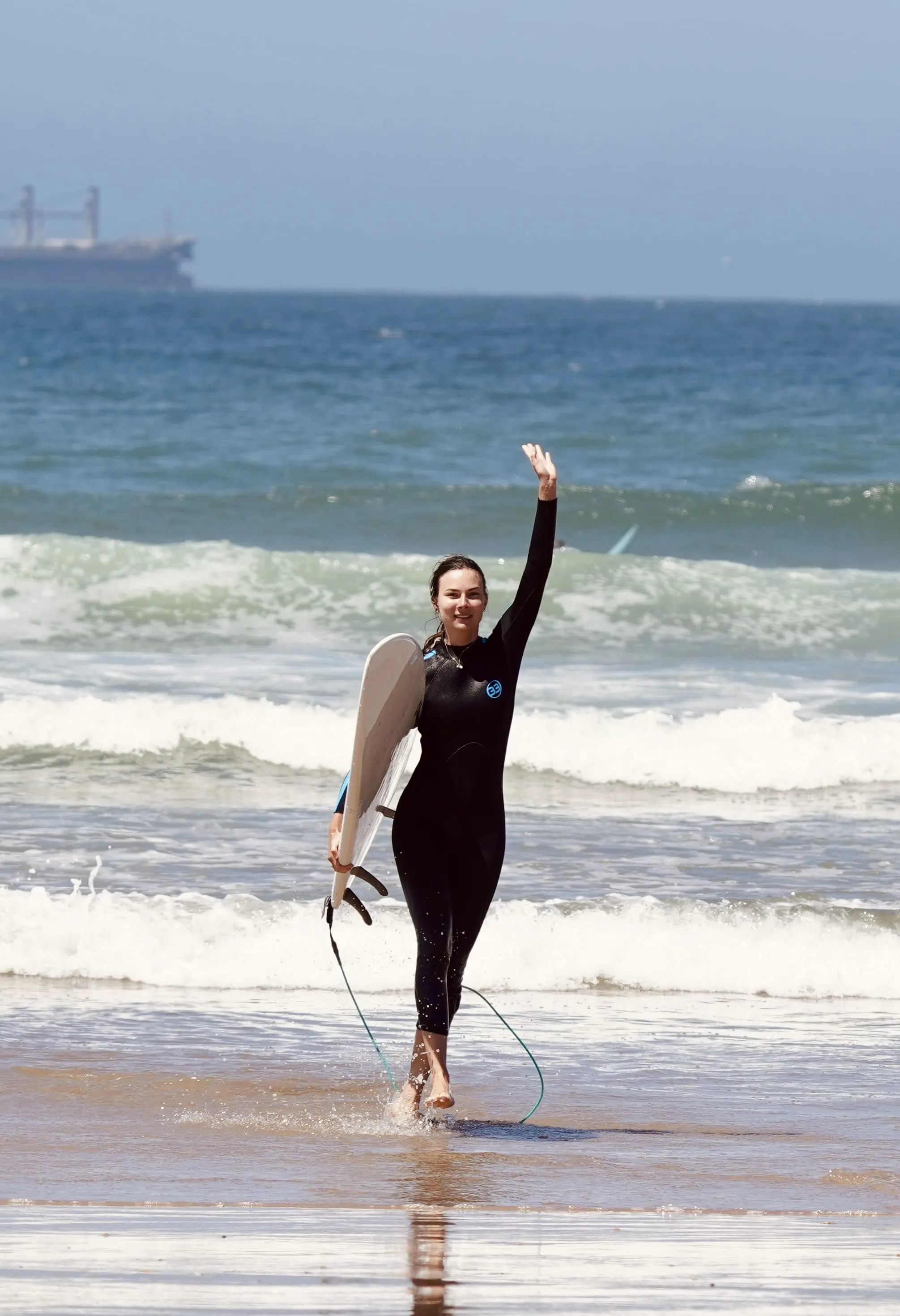 Woman in black wetsuit holding a surfboard, walking on shore with waves and ocean behind her, waving.