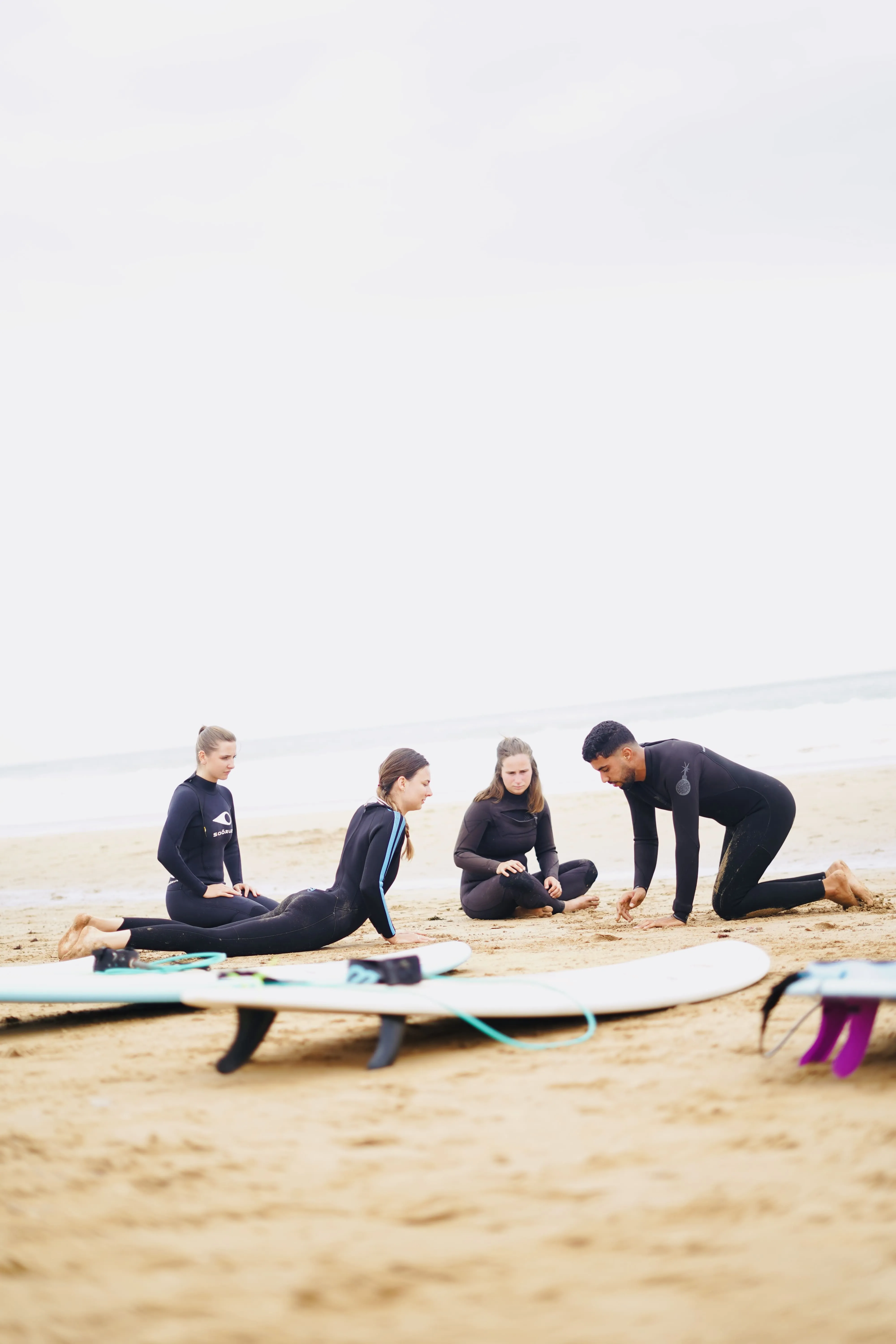 Group of four people in wetsuits on sandy beach with surfboards nearby, preparing for surfing.