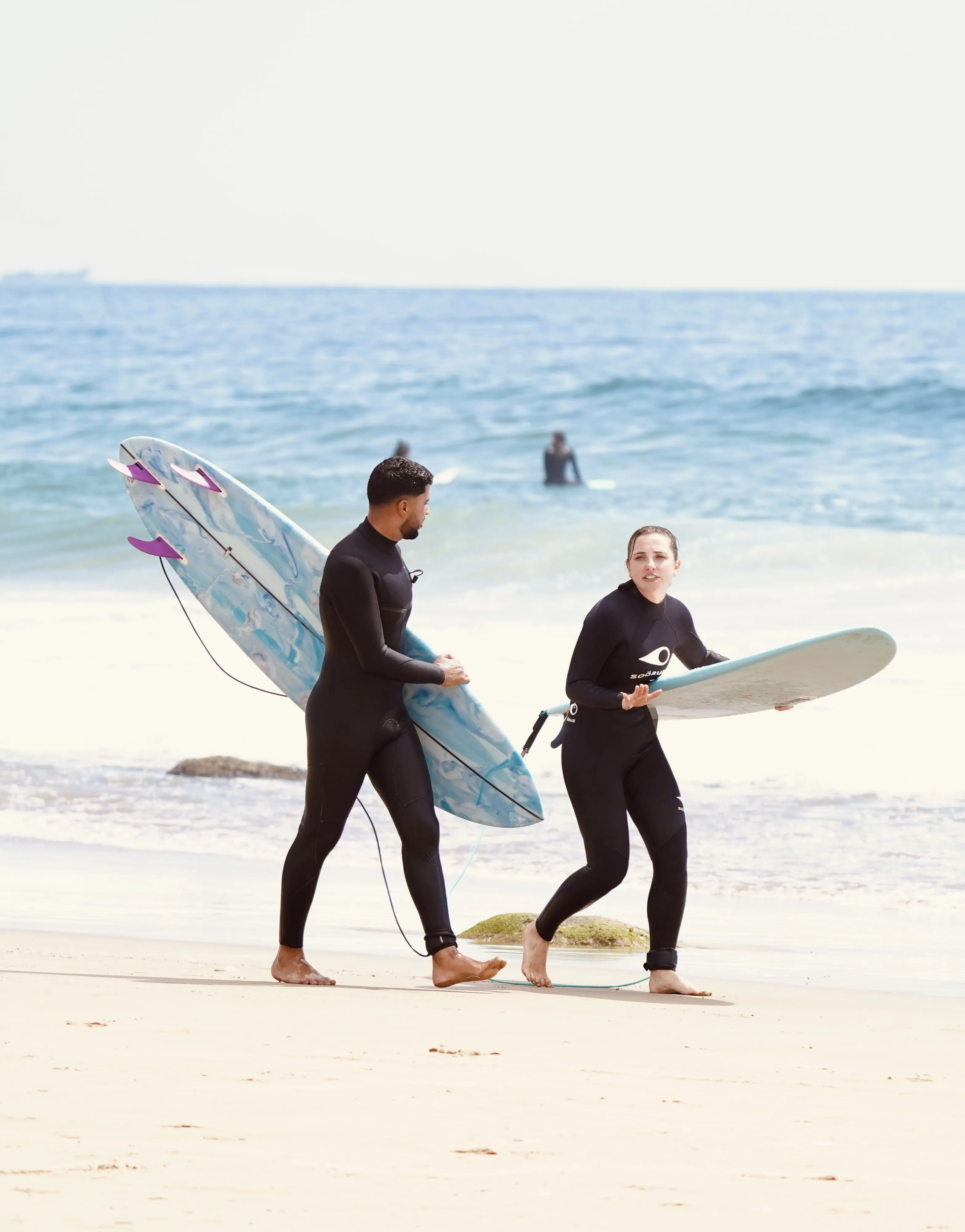 Man and woman in wetsuits walking on beach holding surfboards with ocean waves in background.