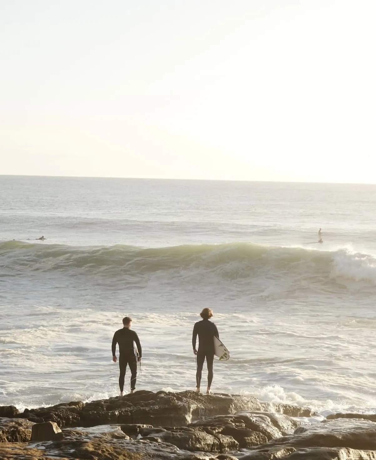 Two surfers in wetsuits standing on rocky shore looking at ocean waves with one holding a surfboard.