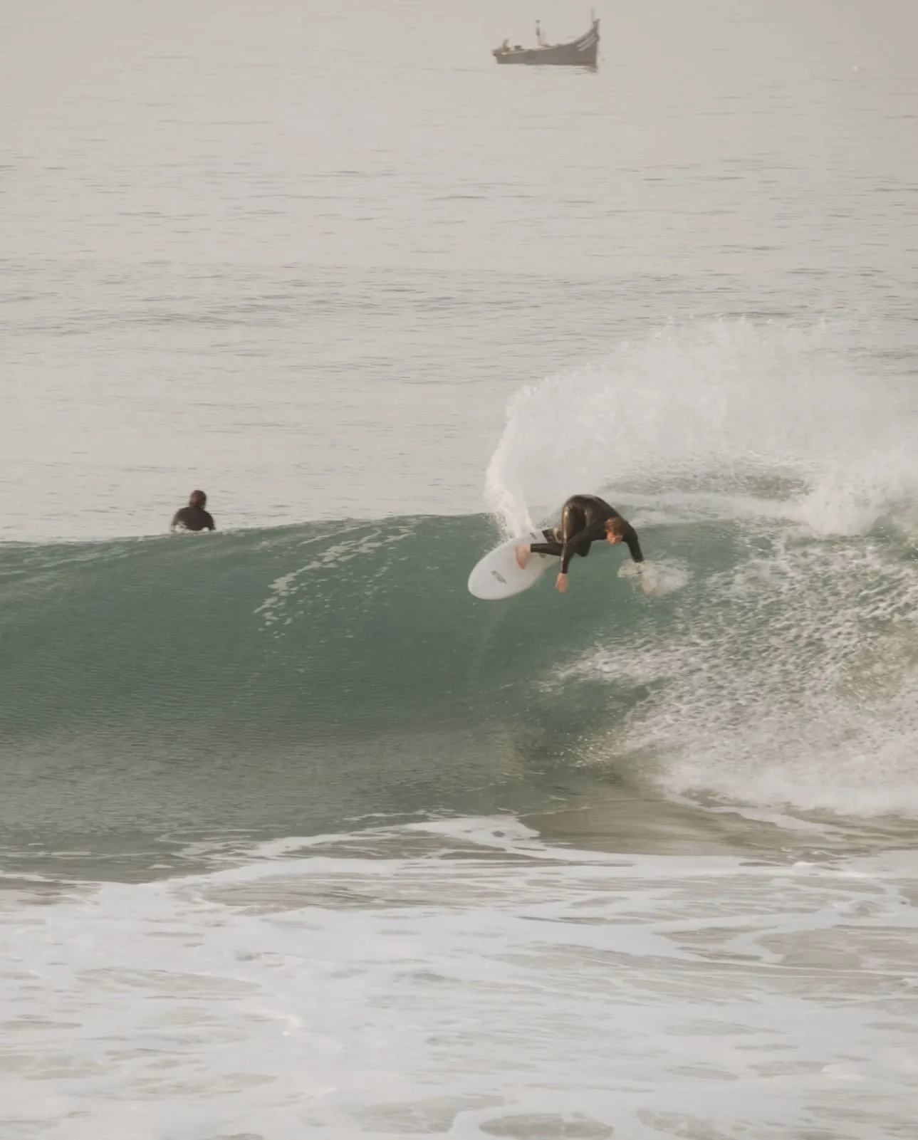 Surfer in a black wetsuit carving a wave with a small fishing boat in the background.