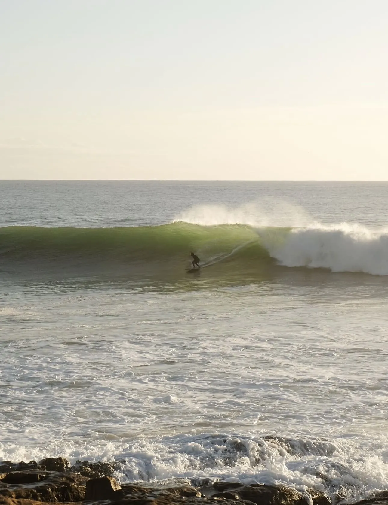 Surfer riding a green wave near rocky shoreline at sunrise or sunset on a calm ocean.