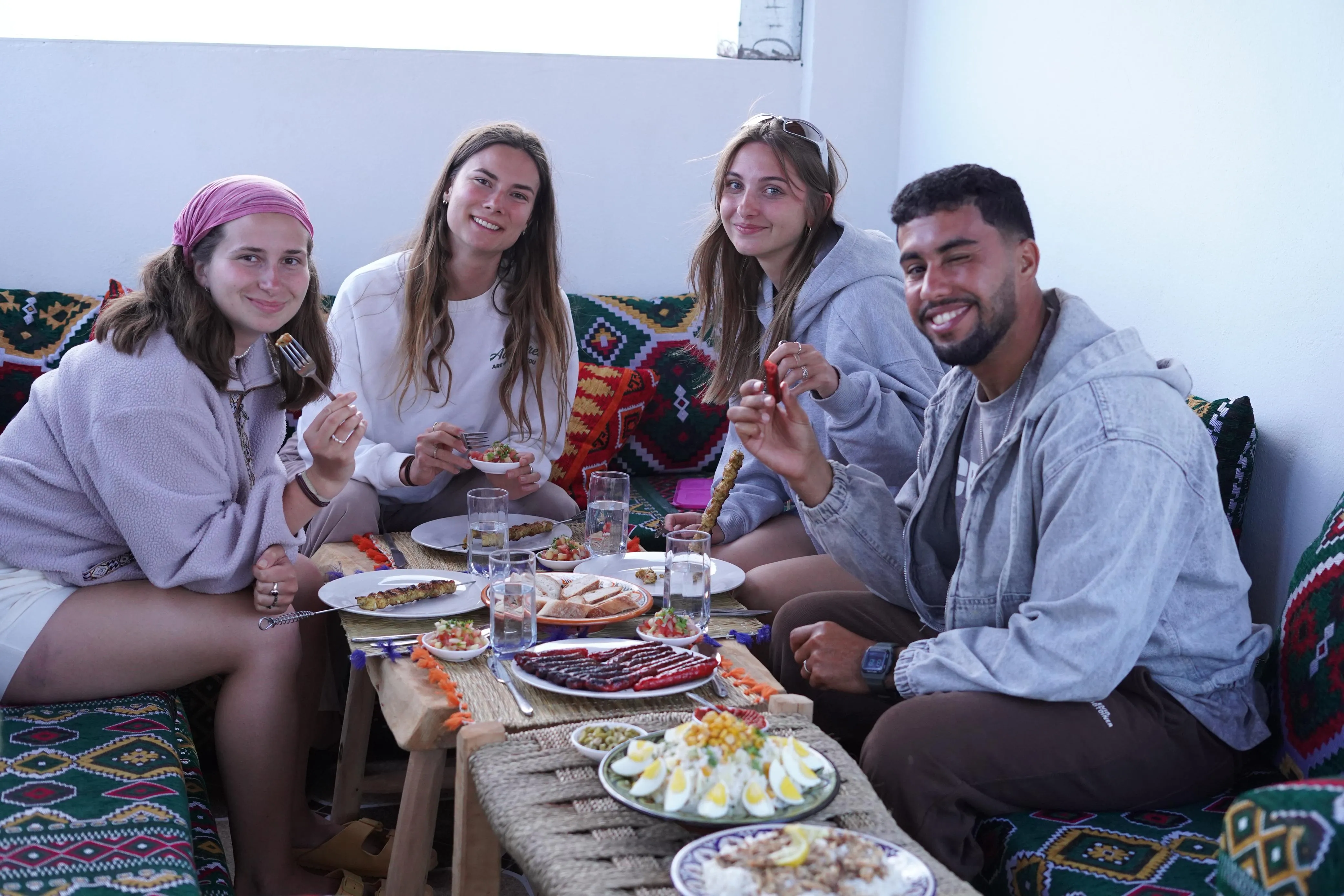 Four friends enjoying a meal with skewers and various dishes around a low table with colorful cushions.