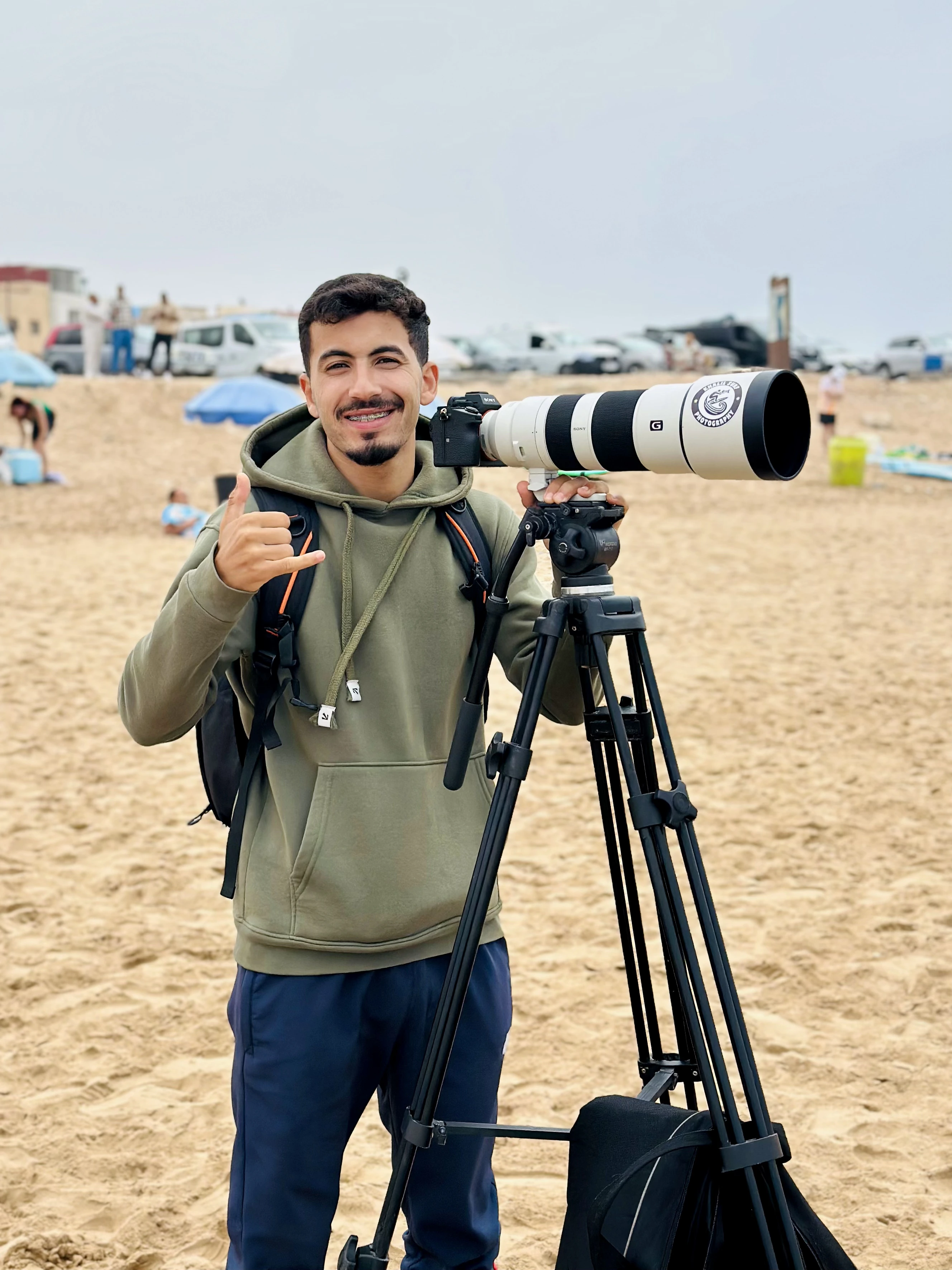 Smiling man in green hoodie giving shaka sign next to camera with large telephoto lens on beach tripod.
