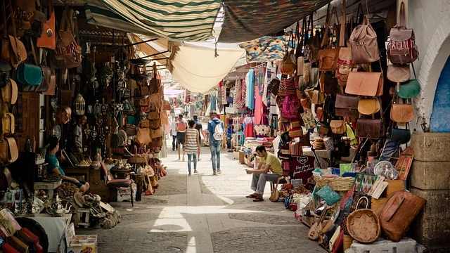 Covered market street with leather goods, baskets, and textiles displayed on both sides and shoppers walking.