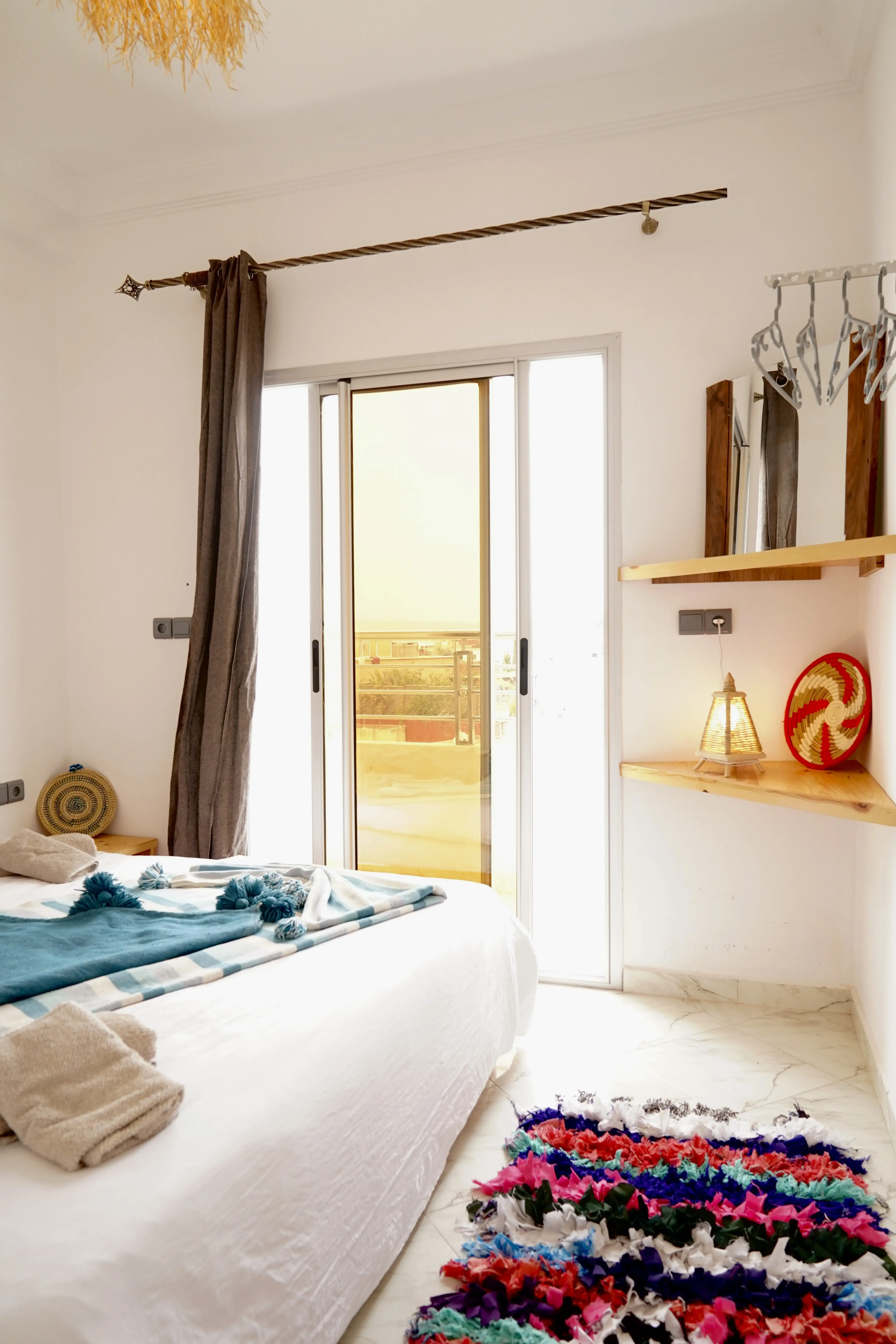 Bright bedroom with white bed, colorful rug, sliding glass door with yellow curtain, and wooden shelves.