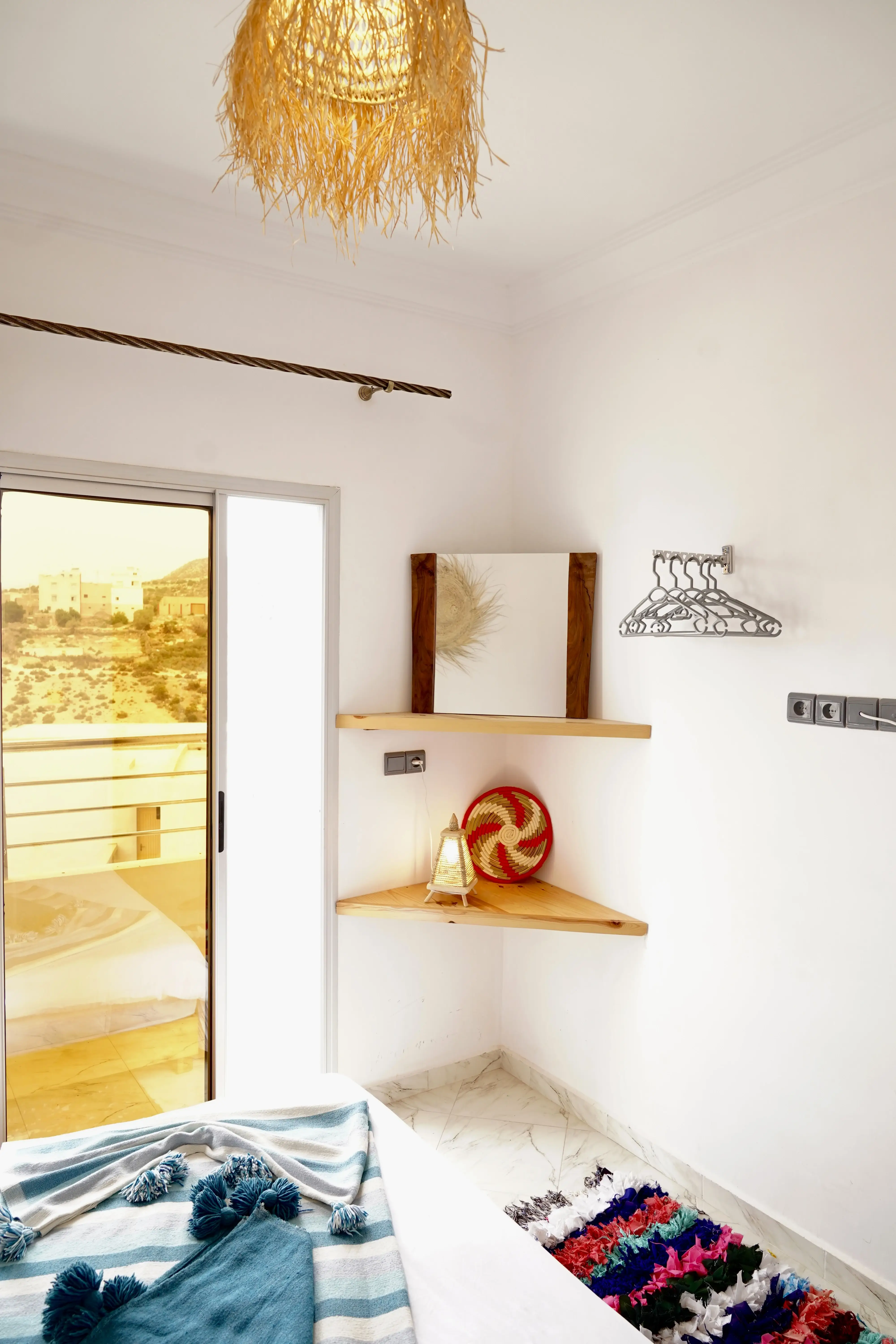 Bright bedroom corner with wooden shelves, hanging silver hangers, striped blanket, and balcony door view.