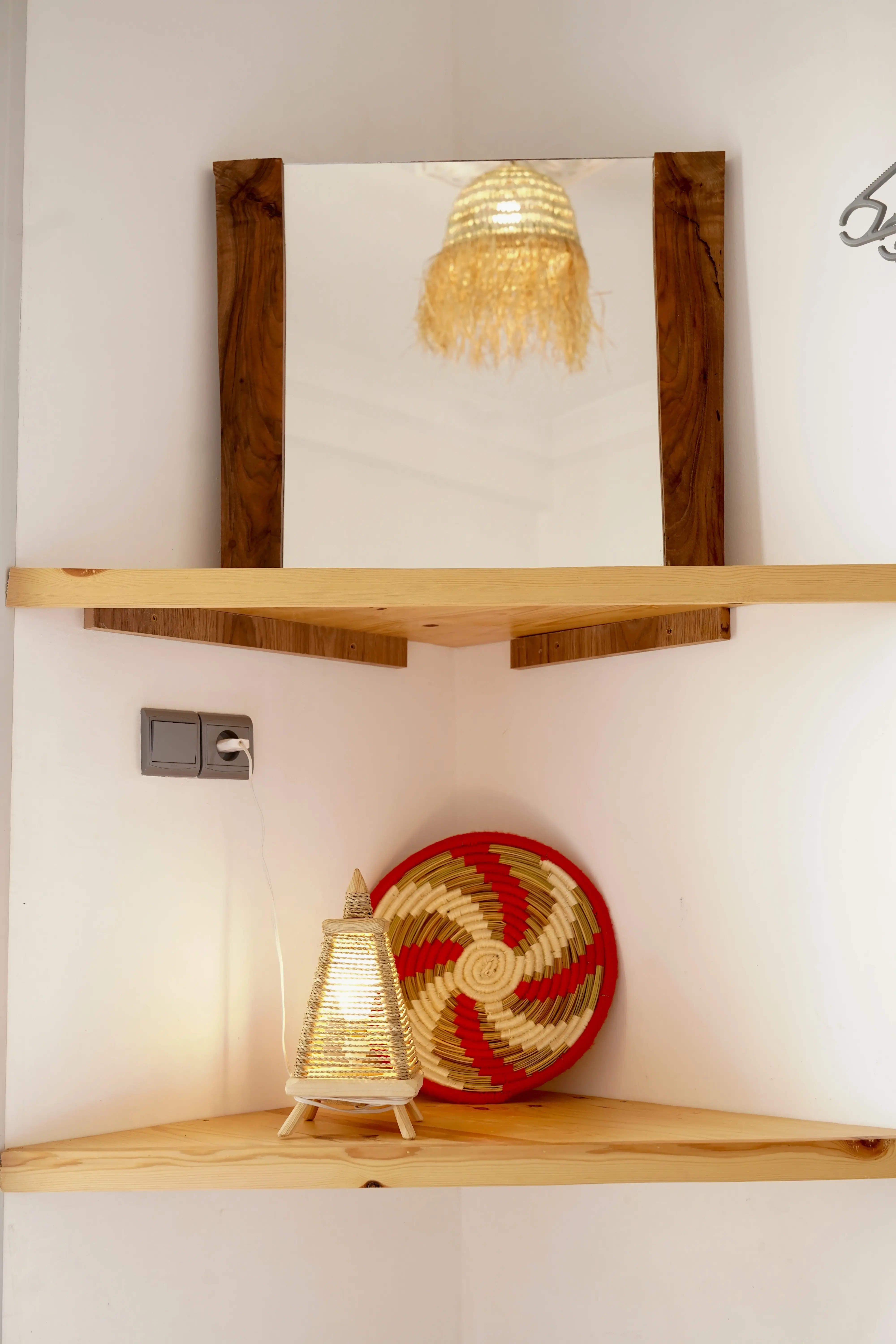 Wood-framed mirror on upper shelf reflects hanging lamp; lamp and wicker basket on lower wooden corner shelf.