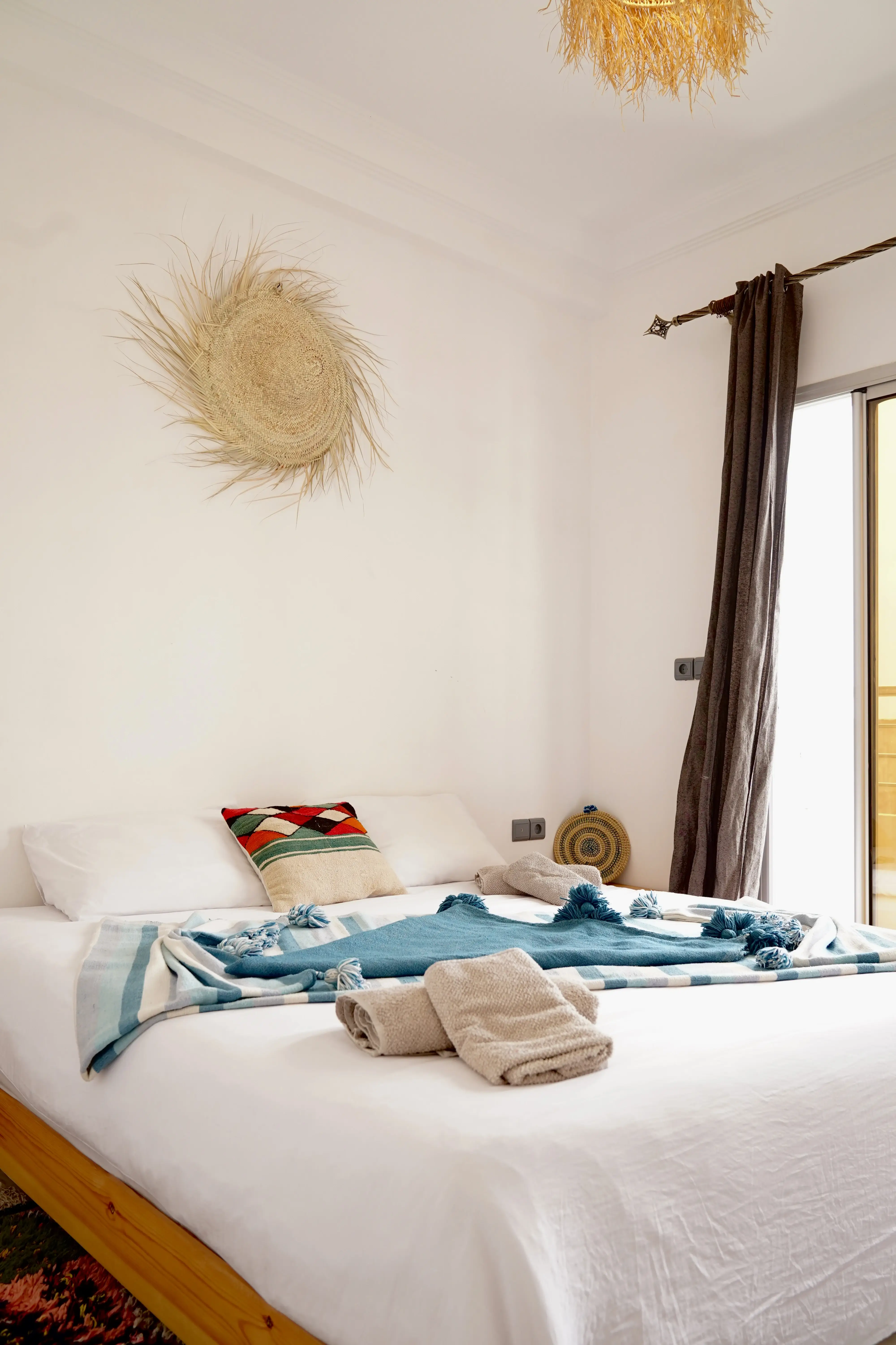 Bedroom with white bedding, patterned pillow, blue throw blanket, rolled towels, and a woven wall decoration.