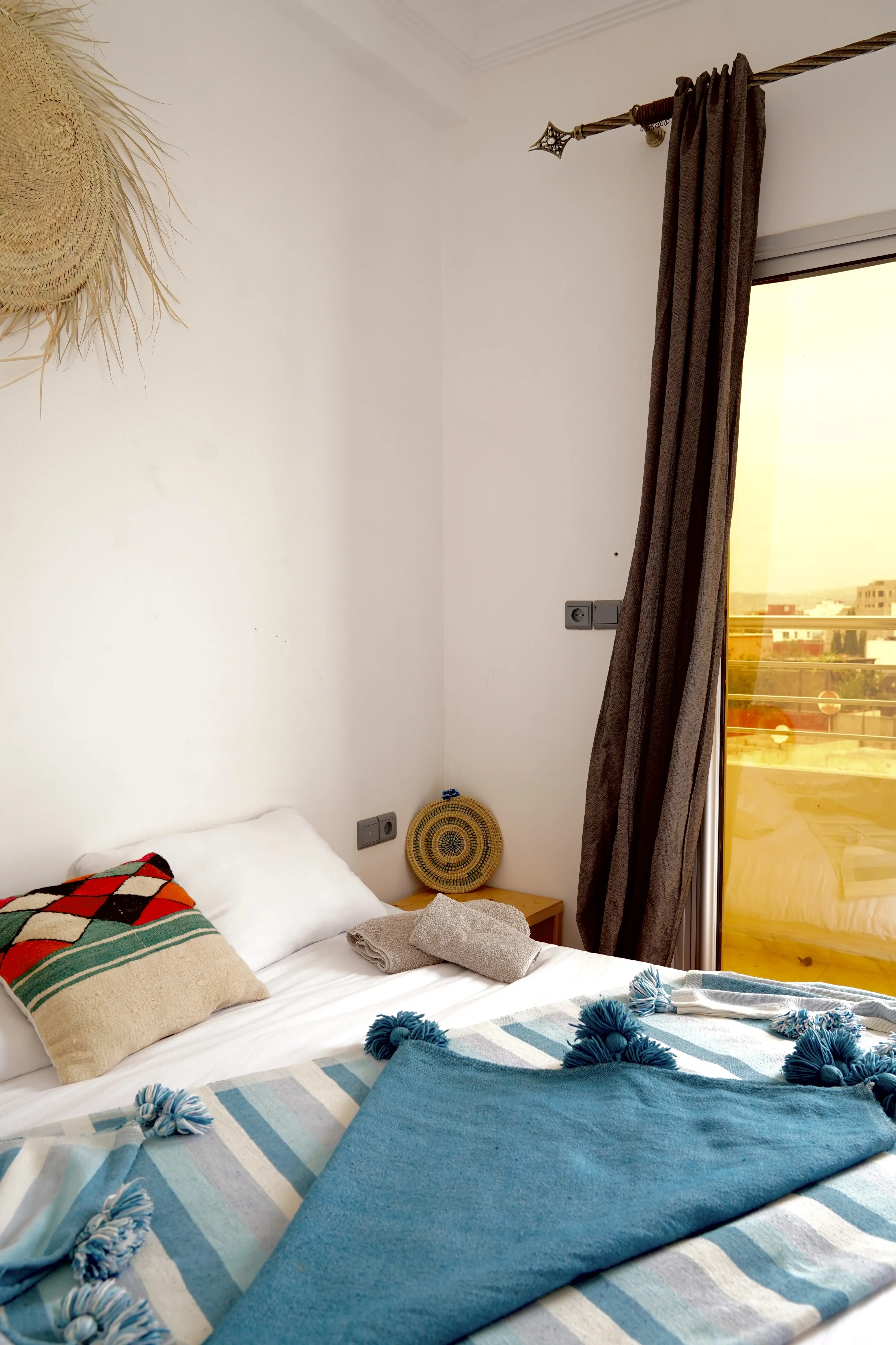 Neat bed with striped blanket, colorful pillow, towels, and dark curtain by sliding glass door.