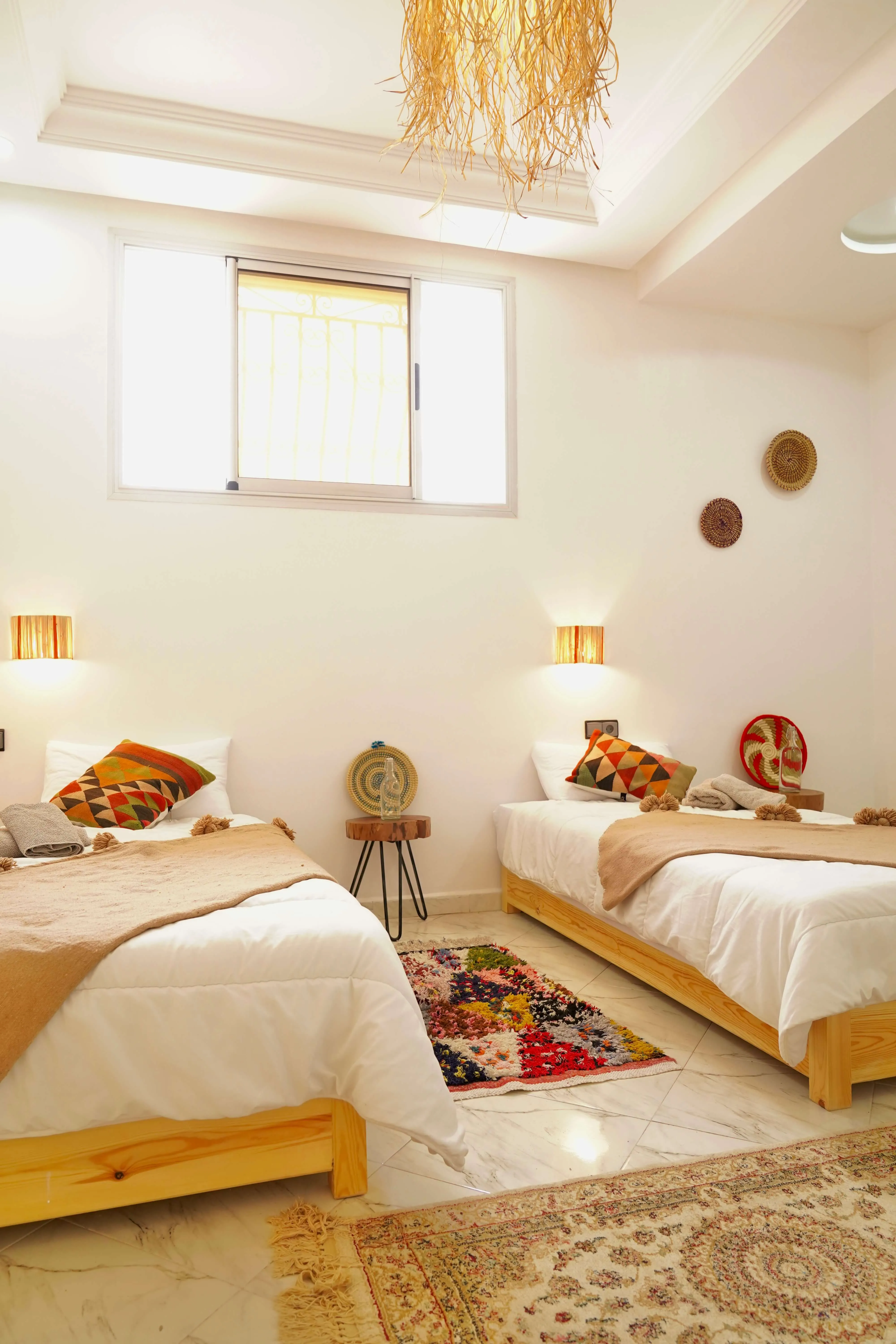 Room with two twin beds with white bedding, beige throws, patterned pillows, and a colorful rug on tile floor.