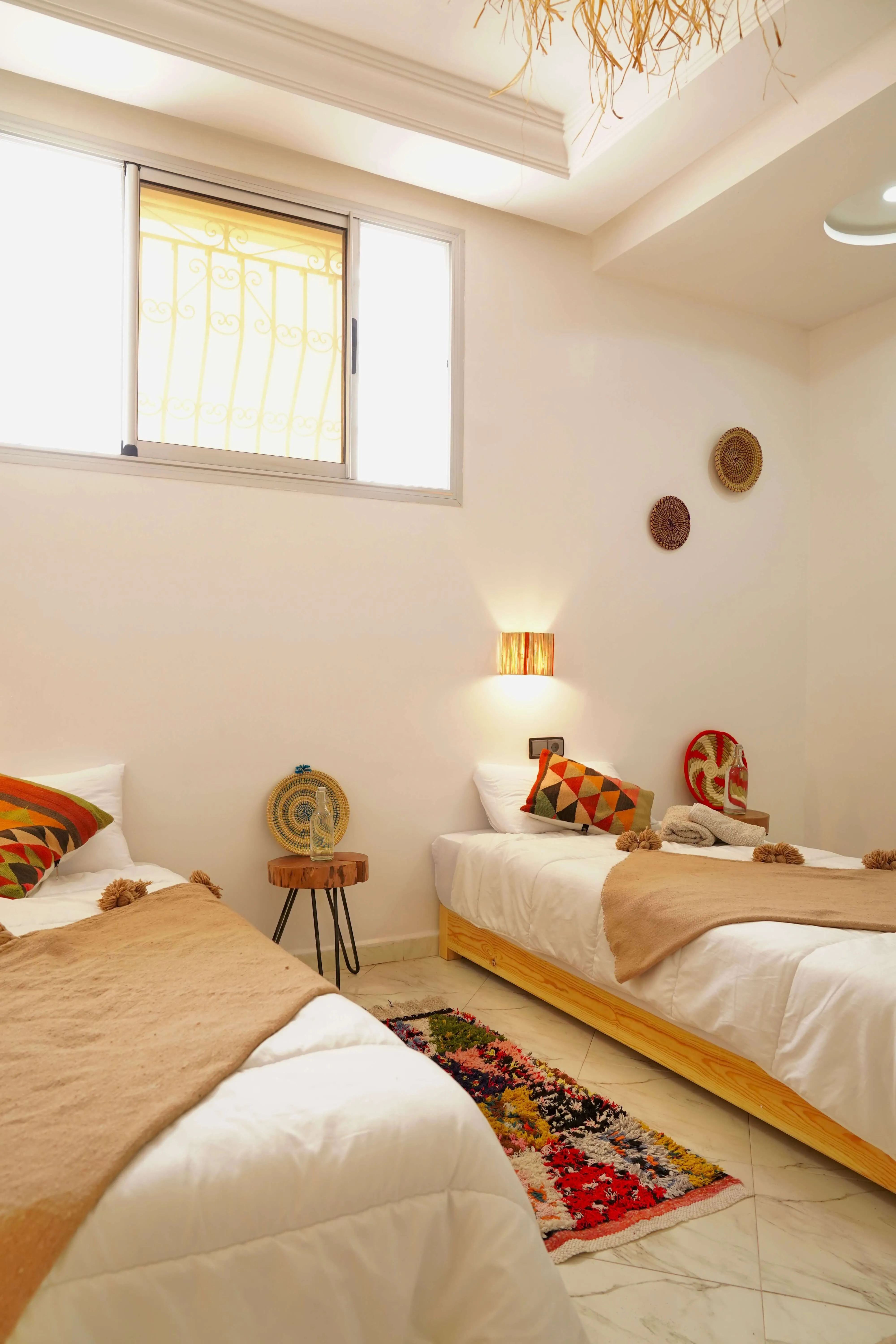 Minimal bedroom with two single beds, colorful pillows, wall lamp, and a small rug on tiled floor.