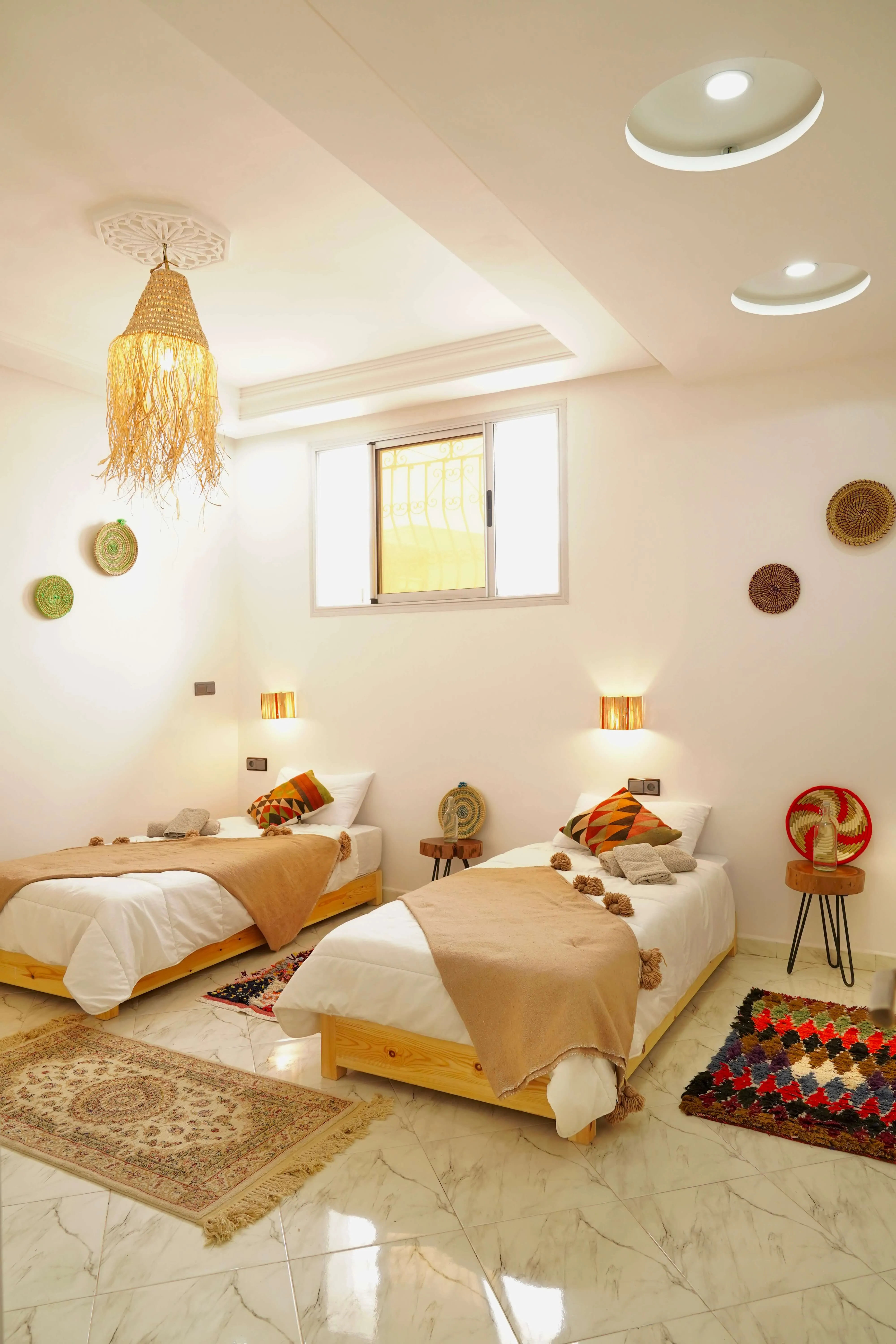 Bright bedroom with two single beds, beige blankets, colorful pillows, rugs, and wall decorations.