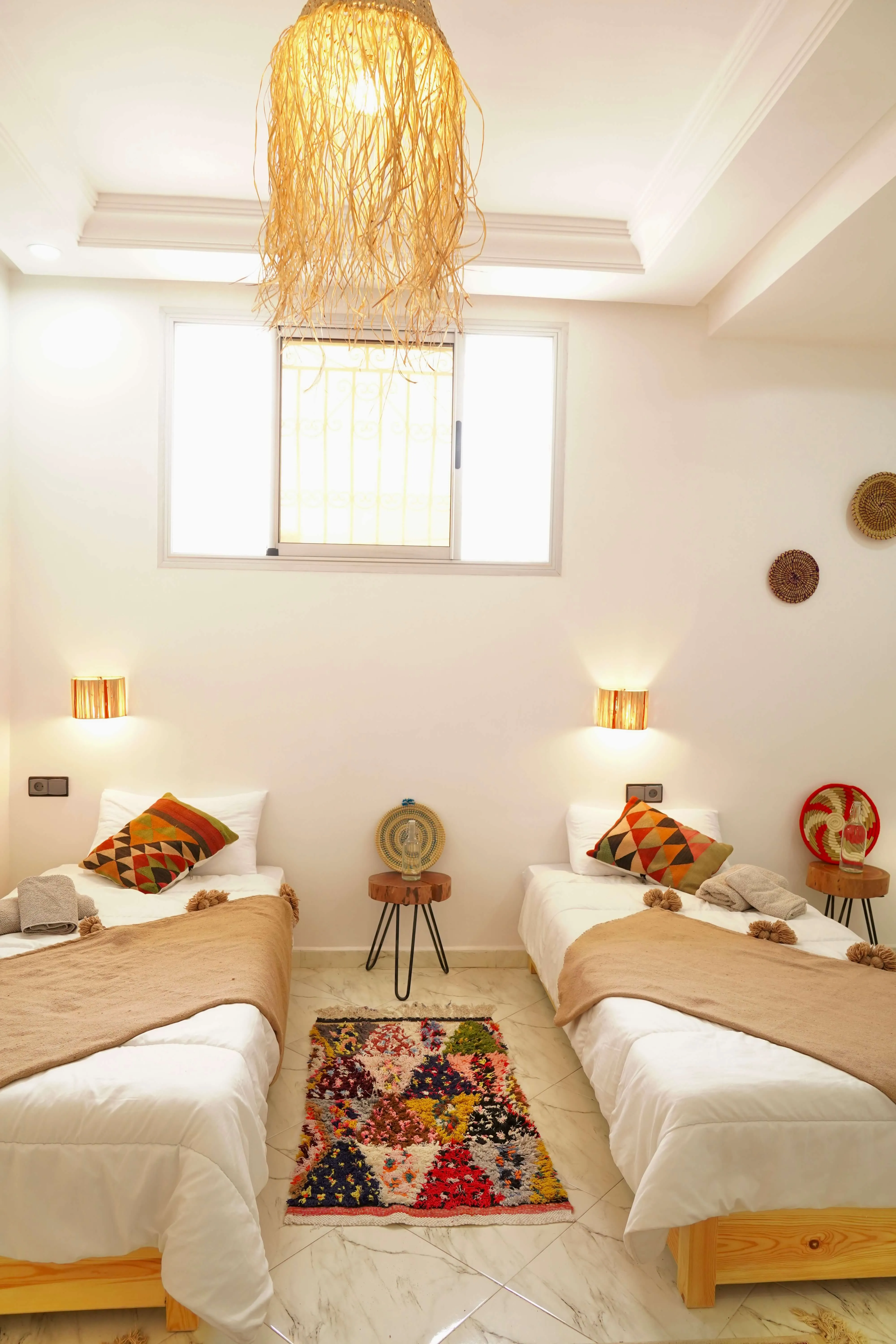 Two single beds with colorful pillows, beige throws, a patterned rug, and a hanging light fixture.