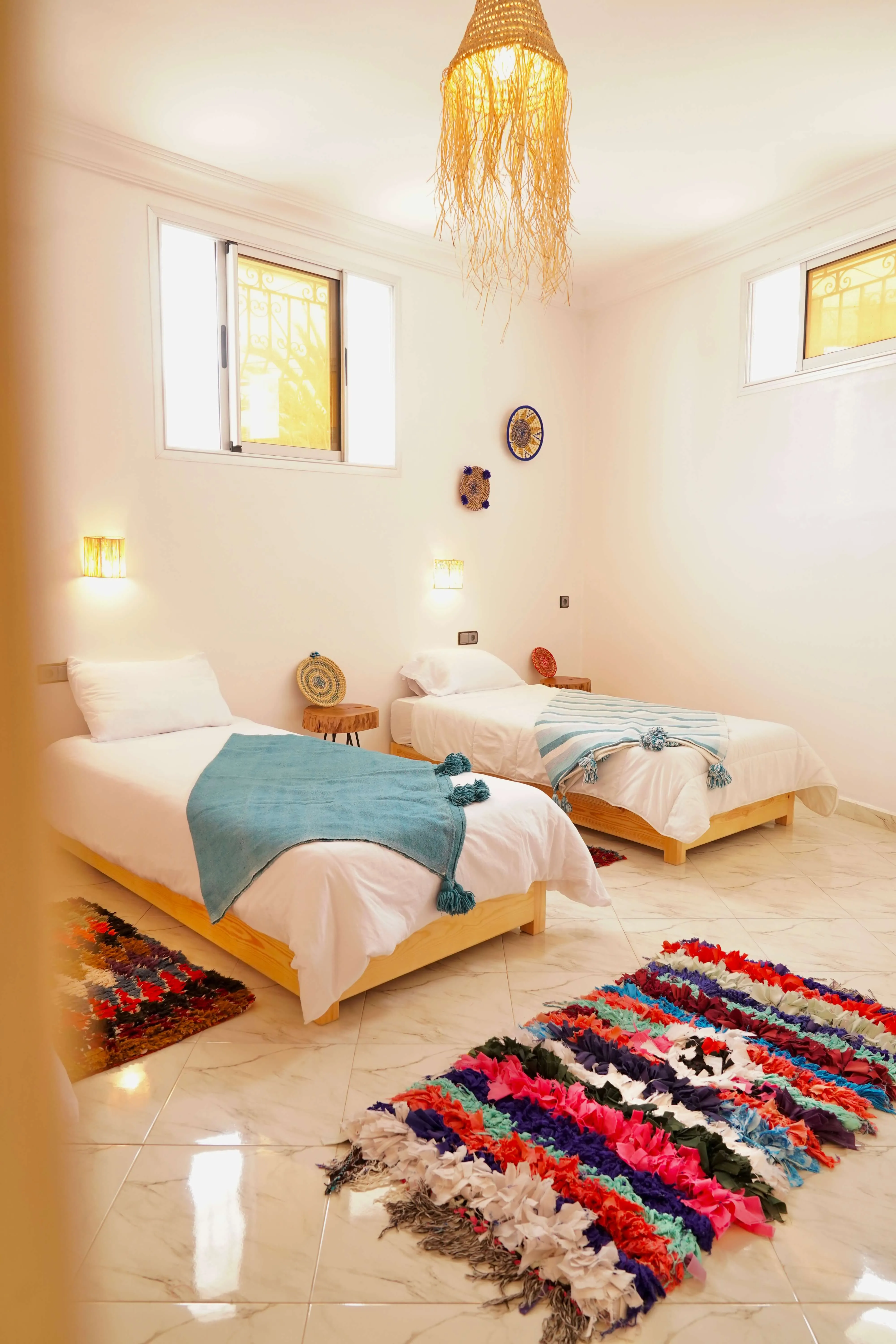 Bright room with two single beds, blue blankets, colorful rugs, wooden stools, and woven lamp.