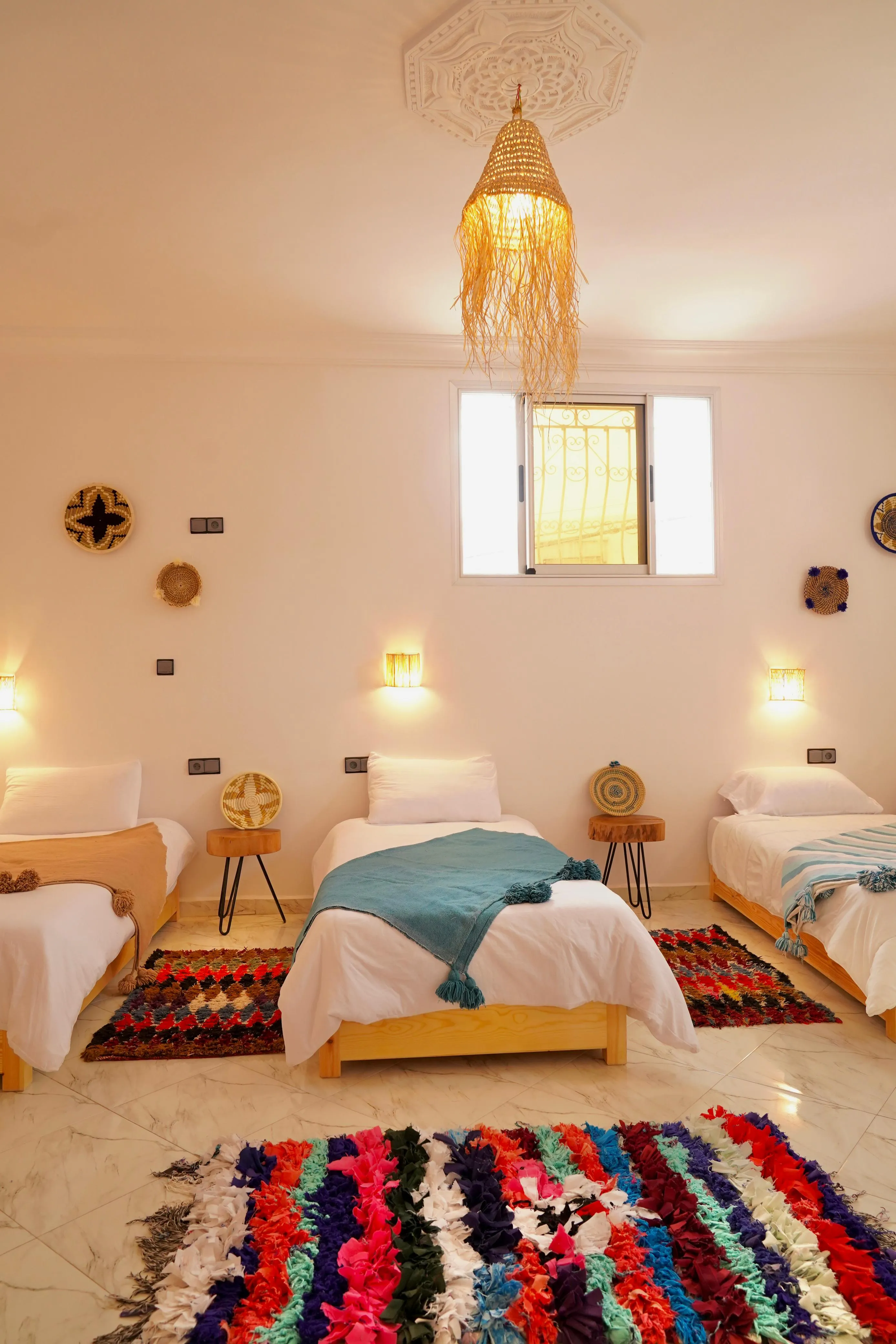 Three single beds with colorful throws in a bright room with woven decor and hanging light fixture.