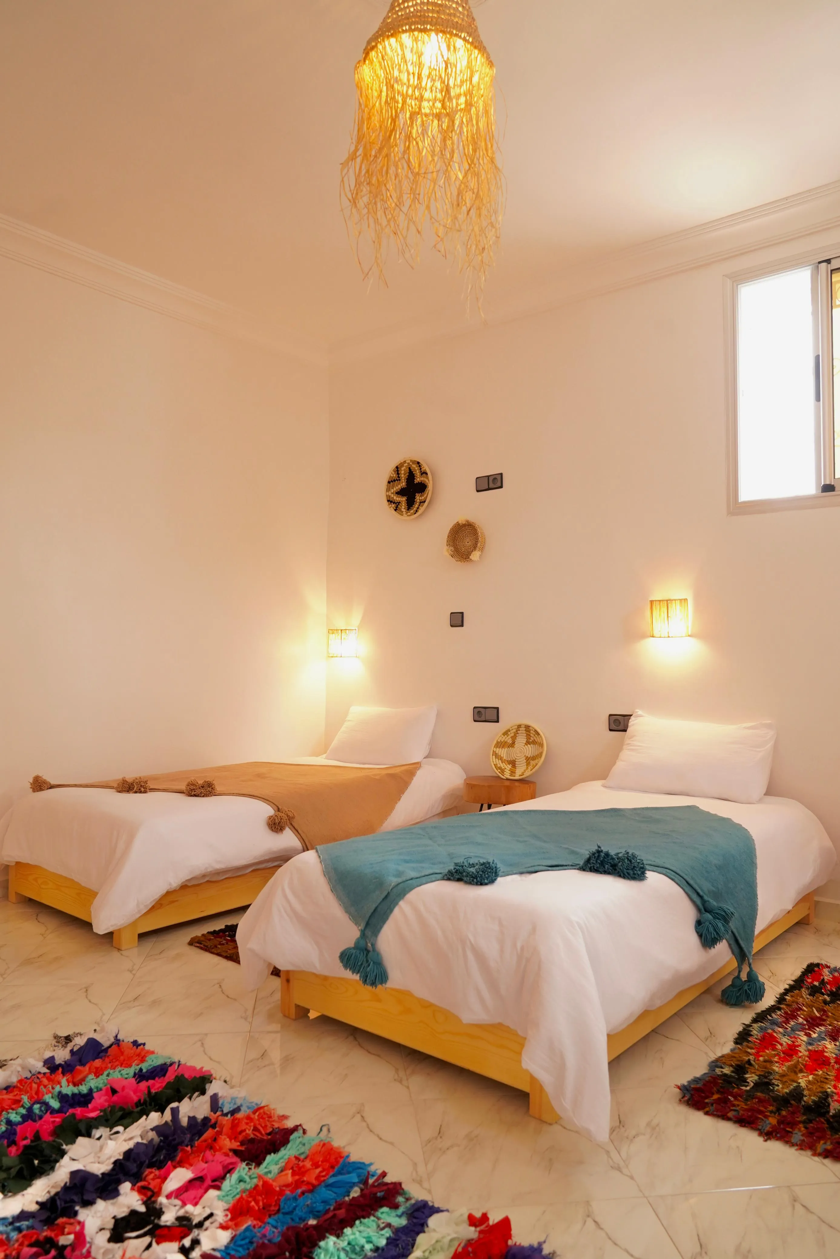 Bedroom with two single beds with white linens, colorful throws, woven wall decor, and a hanging light.