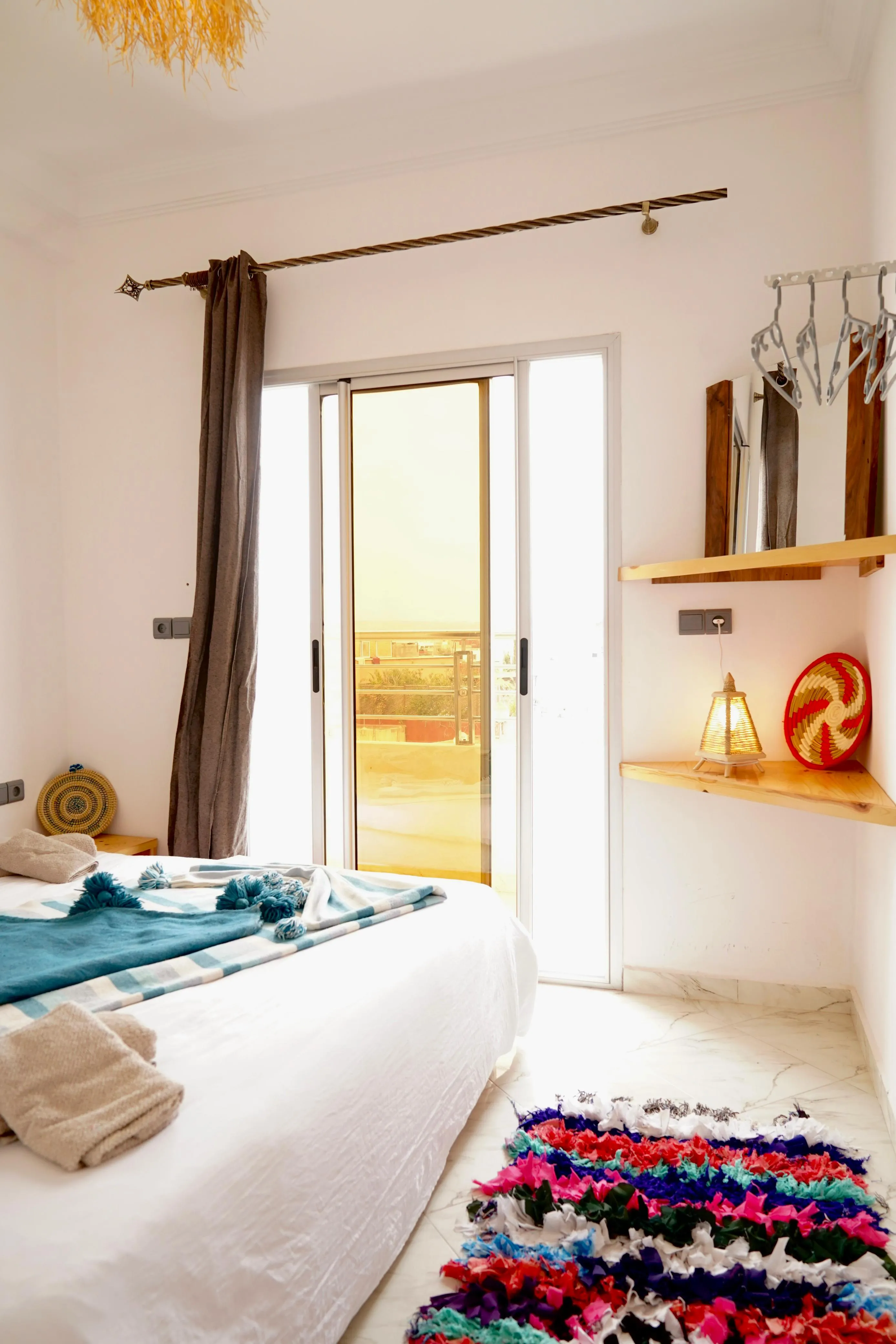 Bright bedroom with bed, blue blanket, colorful rug, balcony door, and wall shelves with lamp and basket.