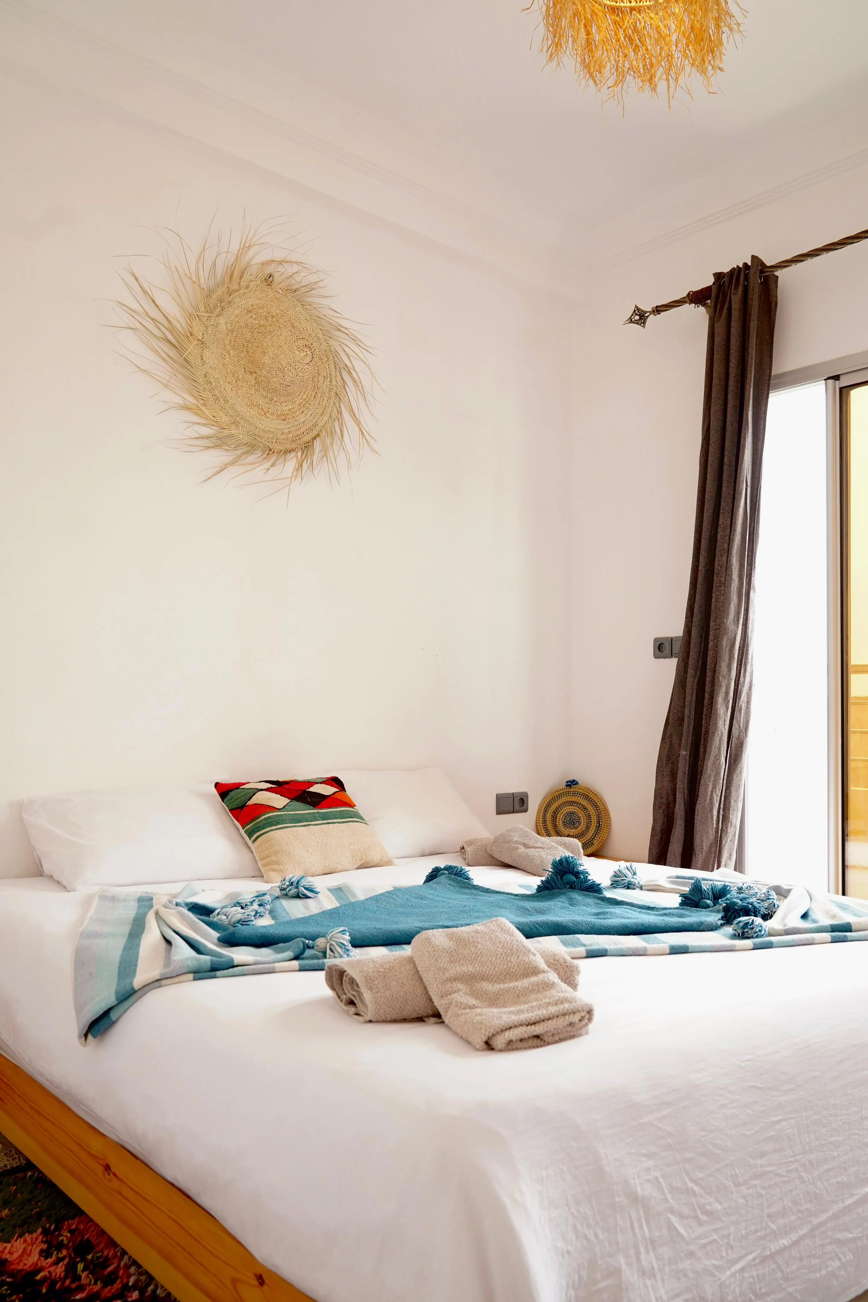 Bright bedroom with white bed, blue throw, folded towels, colorful pillow, and woven wall decor.