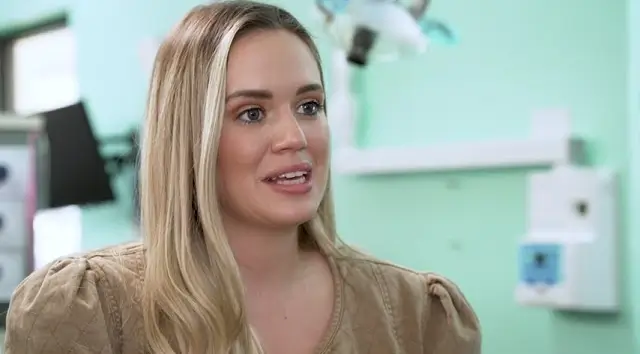 Blonde woman with long hair speaking in a room with medical equipment and turquoise walls.