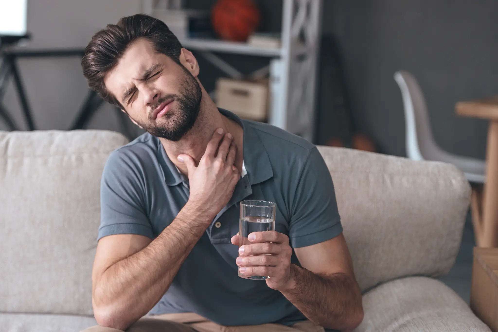Man sitting on a couch holding a glass of water and clutching his throat in discomfort.