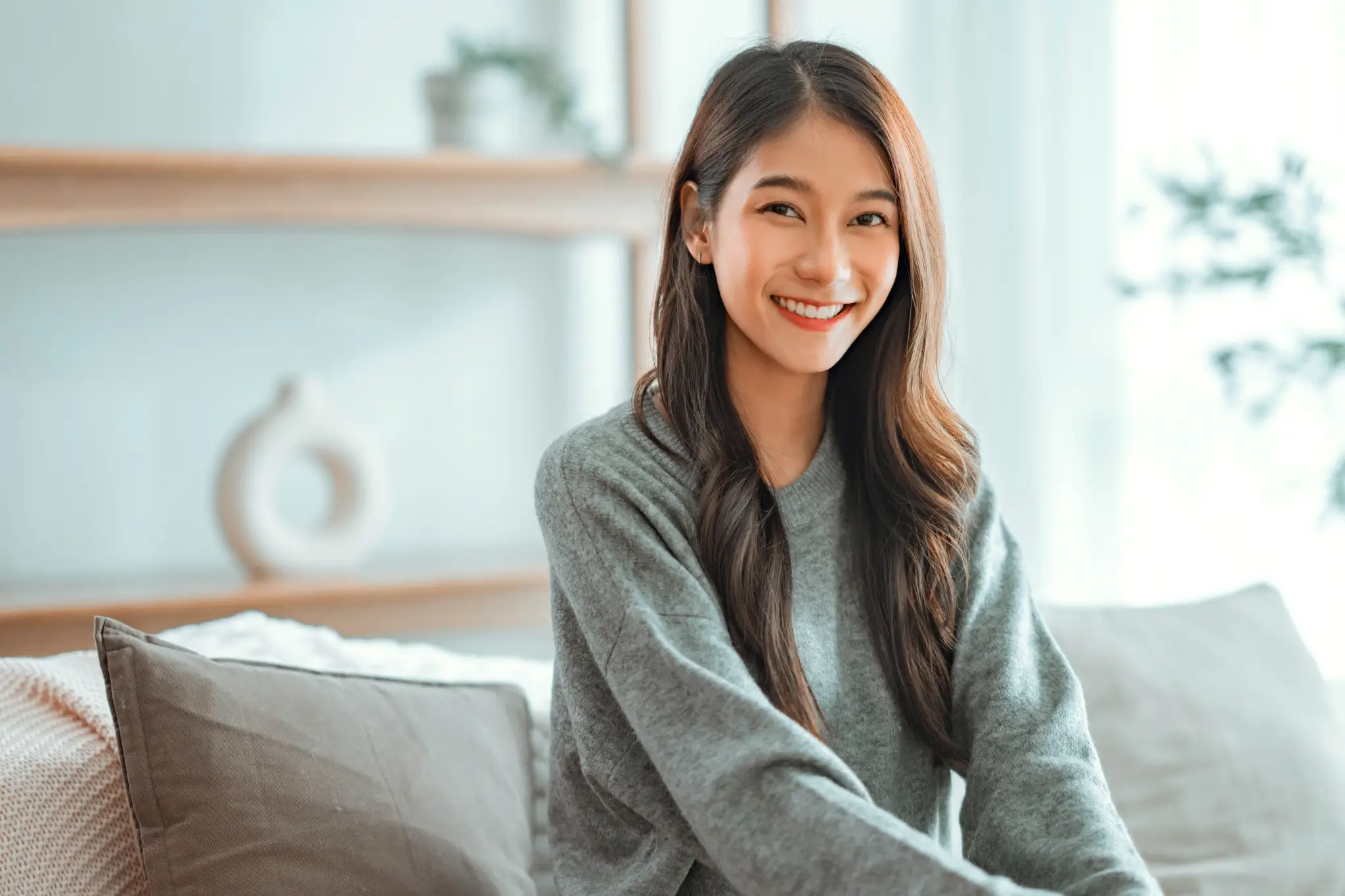 Smiling young woman with long hair wearing a gray sweater sitting on a couch in a bright, modern living room.