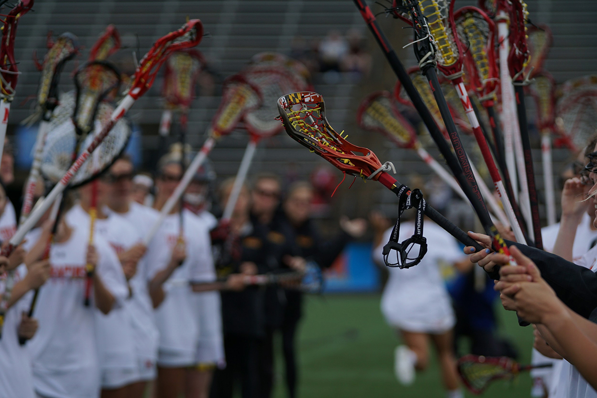 Group of lacrosse players in white uniforms raising sticks with a close-up of one stick with Maryland flag pattern.