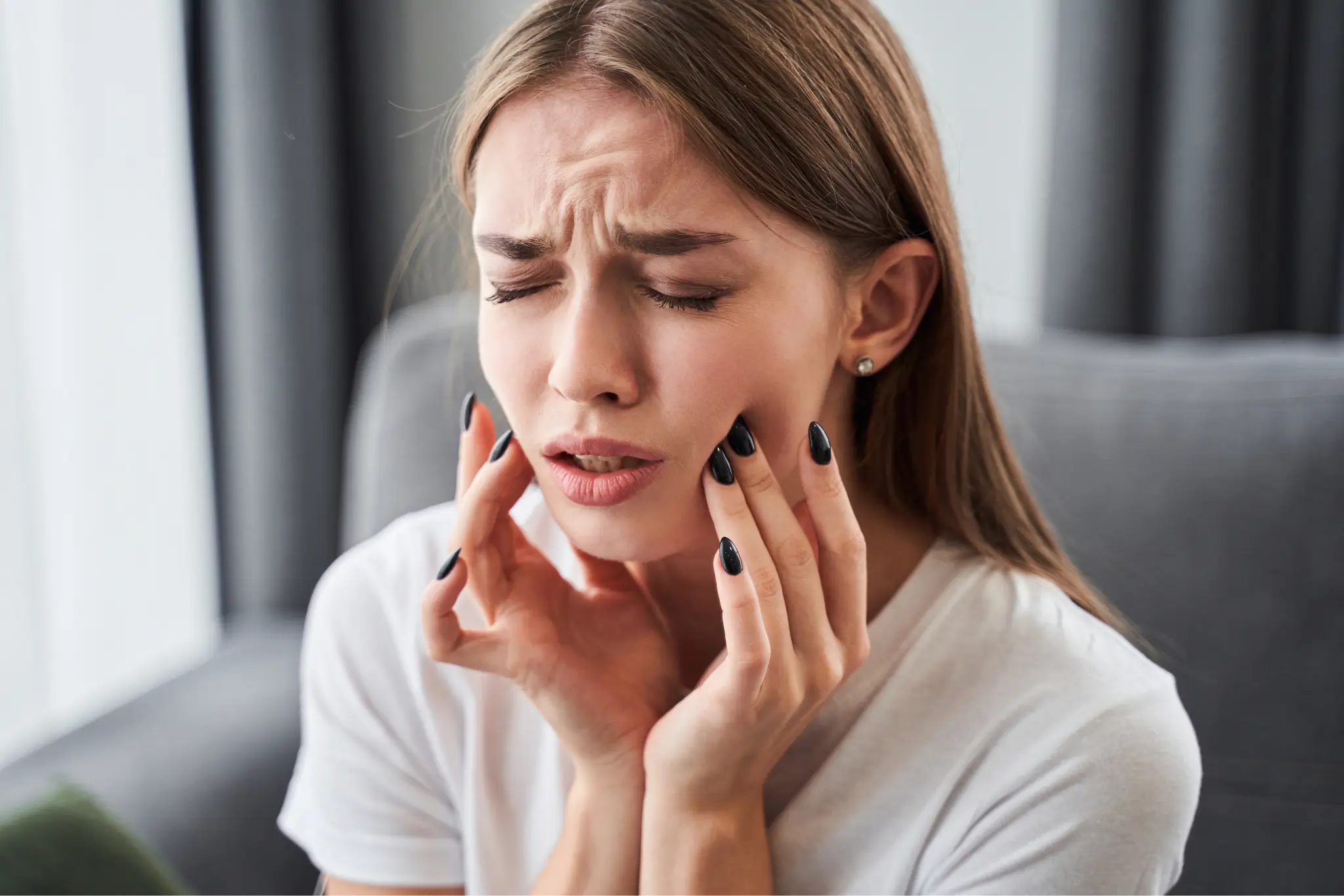 Woman in white shirt holding her jaw with eyes closed, expressing toothache or facial pain.
