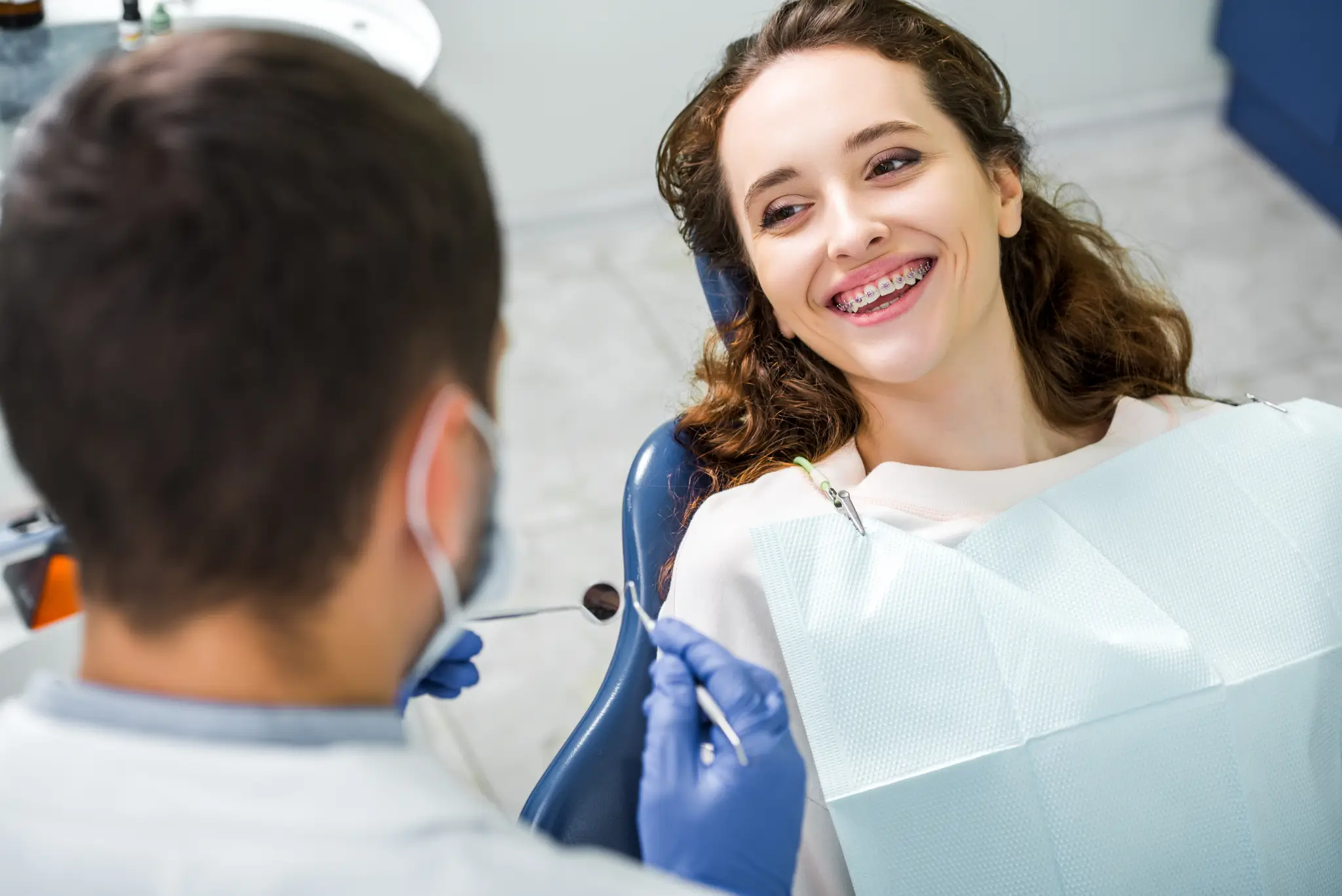 Smiling woman with dental braces sitting in a dentist chair while dentist wearing blue gloves holds dental tools.