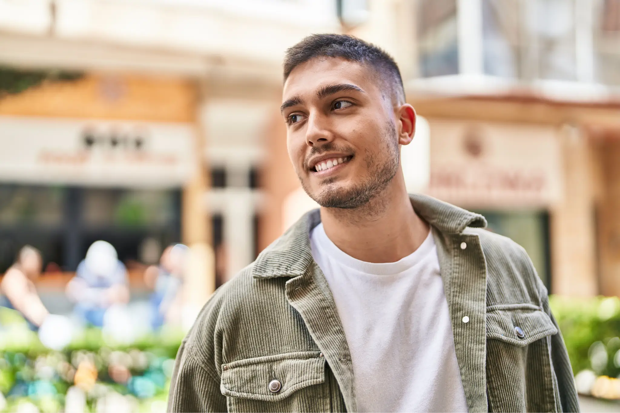 Smiling young man with short hair wearing a white t-shirt and green jacket standing outdoors in an urban setting.