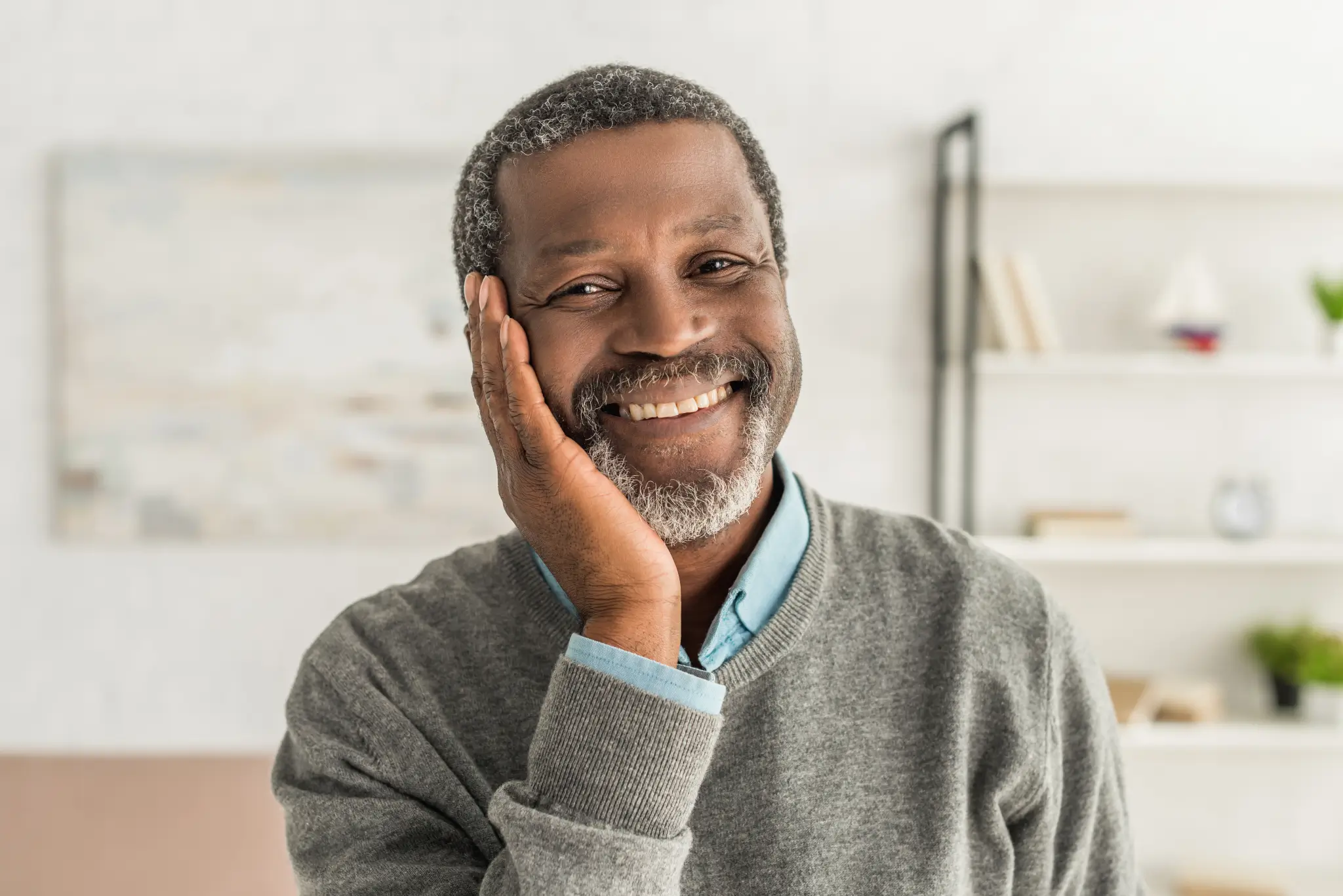 Smiling older man with gray hair and beard resting head on hand in a cozy living room.