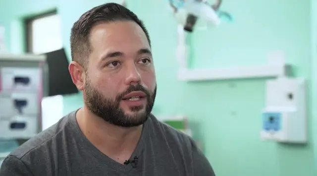 Man with short dark hair and beard wearing a gray shirt, speaking indoors with a blurred medical or dental equipment background.