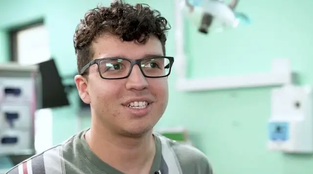 Young man with curly hair and glasses smiling in a room with light green walls and medical equipment.