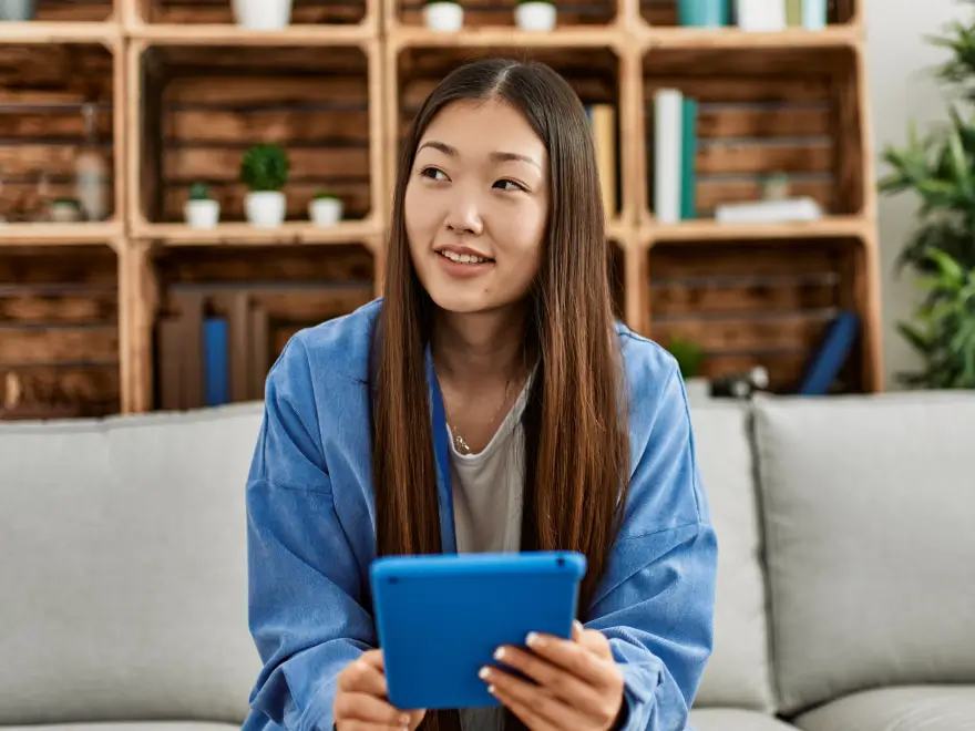 Young woman with long straight hair sitting on couch holding a blue tablet and looking to the side.