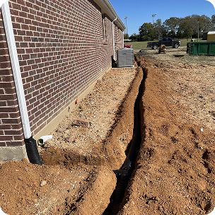 Trench dug alongside a brick building with drainage pipes and gravel near HVAC units outdoors.