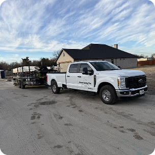 White pickup truck parked in a driveway with a trailer carrying lawn equipment, a house and cloudy sky behind.