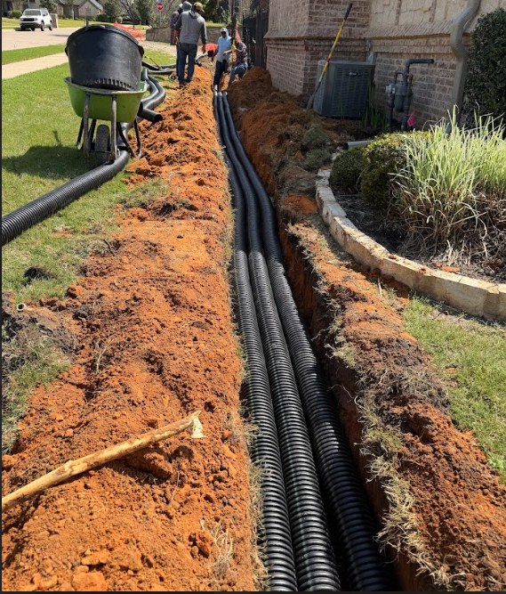 Trench next to house with black corrugated pipes laid inside and workers in the background on a sunny day.