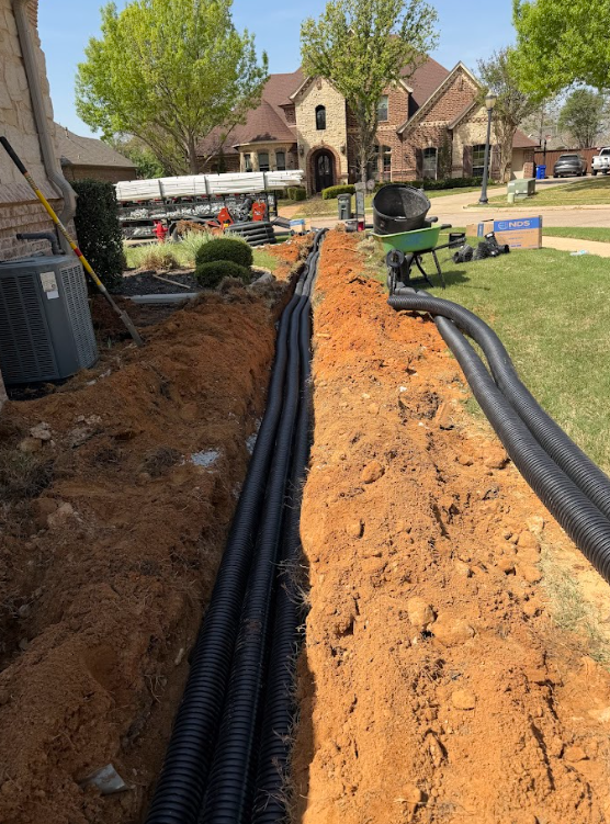 Trench with black drainage pipes being installed in front yard of a suburban brick house on a sunny day.