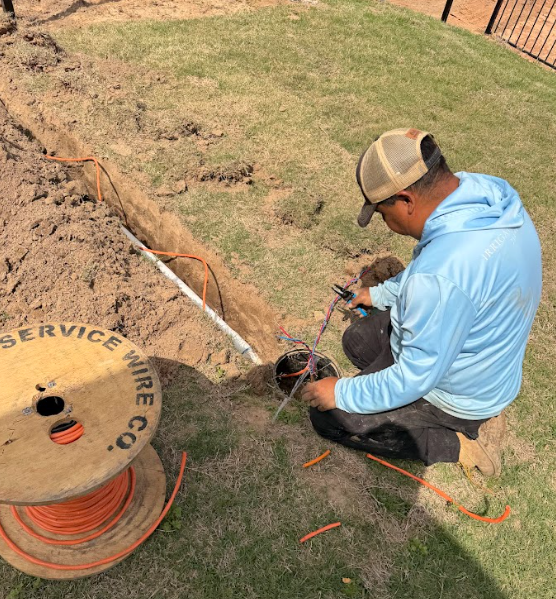Worker kneels on grass handling multicolored wires near a trench and an orange wire spool during outdoor wiring work.